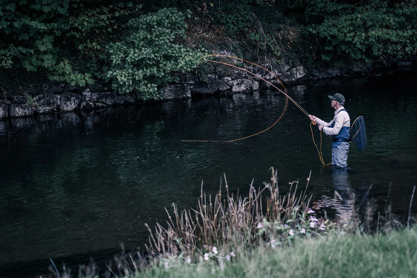 Fly fisherman wading in a forest-lined river with a fly rod and landing net