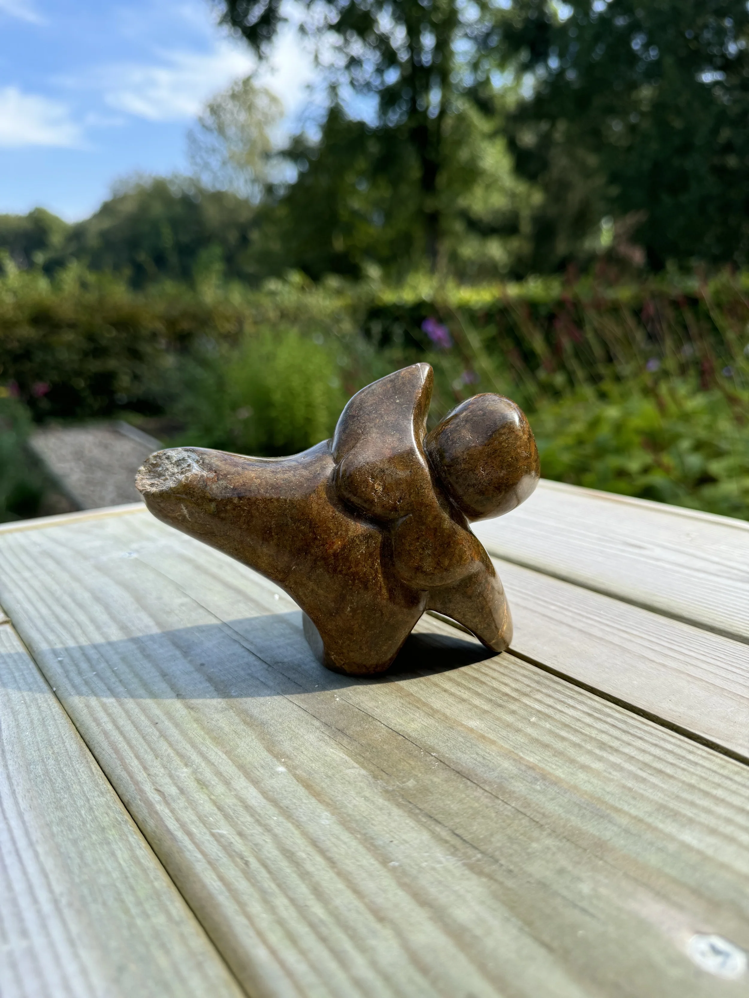 Beeld van bruin speksteen, een figuur die op een been en arm steunt de andere arm en been steken in de lucht.
