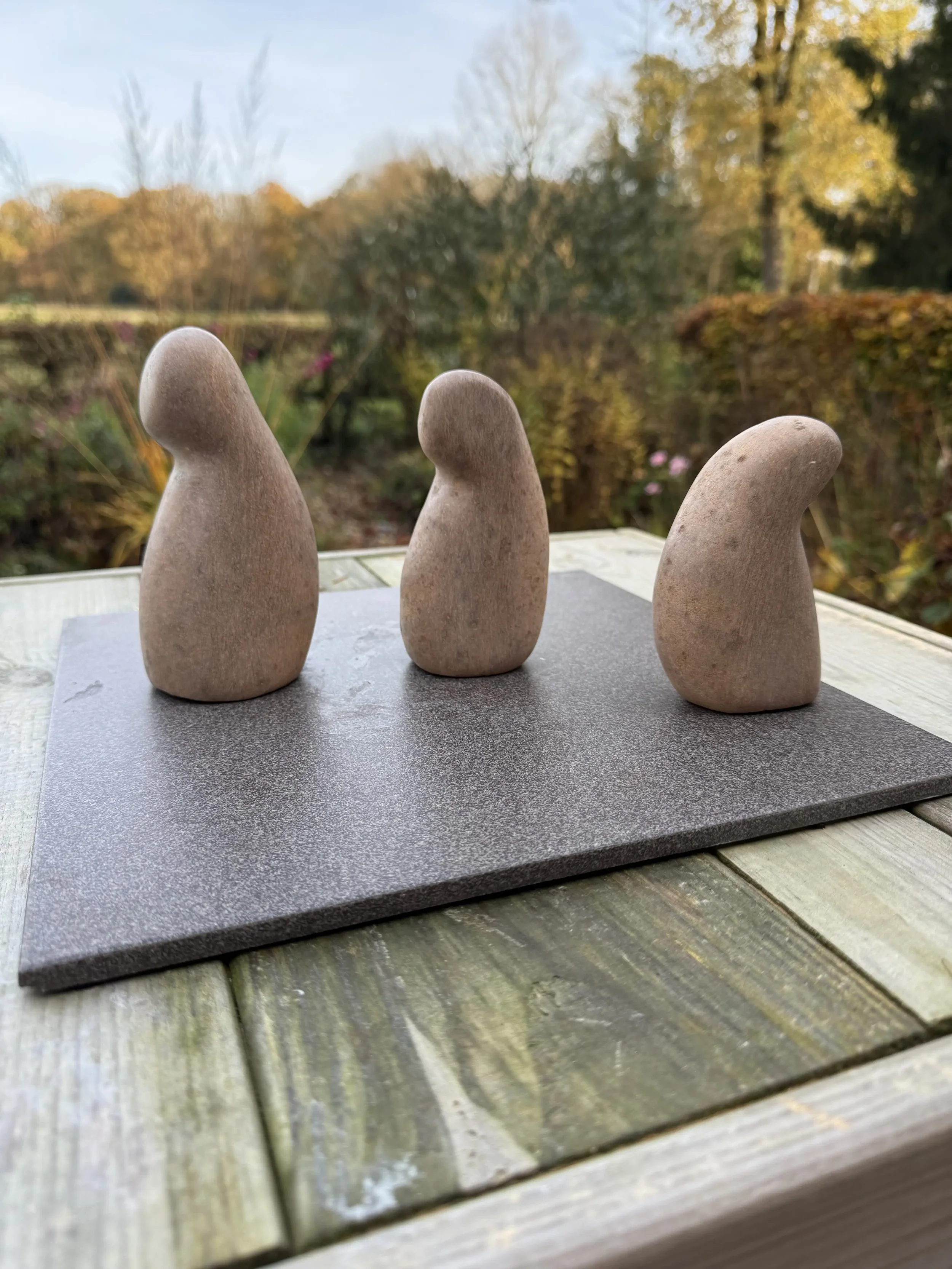 De drie figuren van Familie van bruin speksteen ongepolijst staand op een dunne grijze tegel op een houten beeldhouwbok