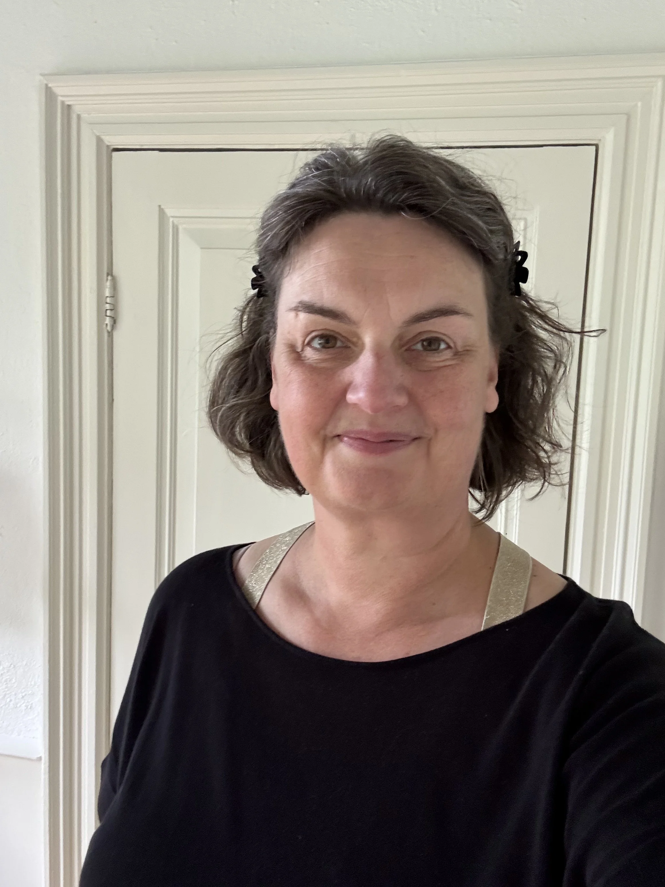 Close-up van een glimlachende vrouw met kort bruin haar met een haarclip, gekleed in een zwarte top met een beige of goudkleurige band, voor een witte deur met decoratieve rand.