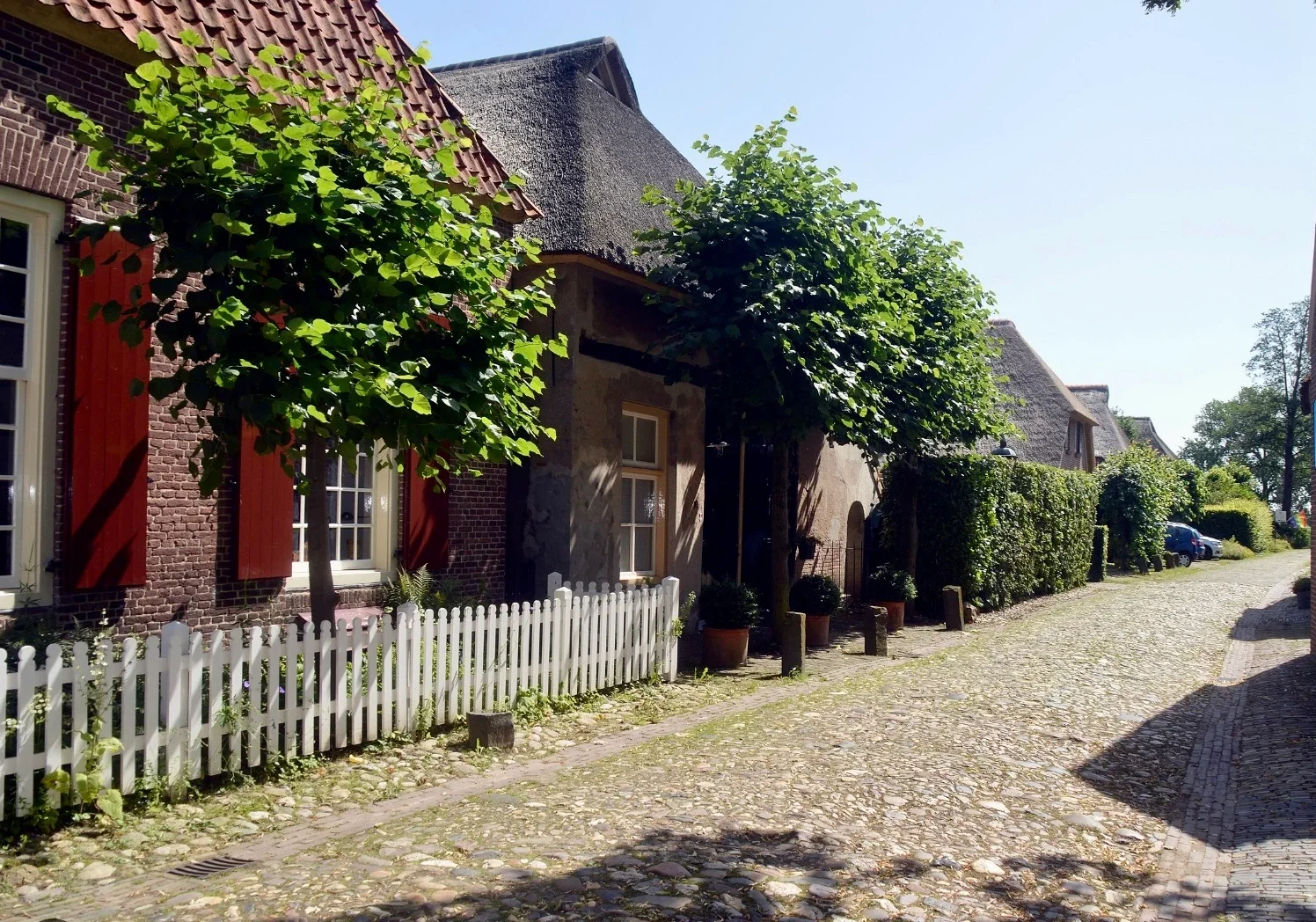 Een rustige, kasseienstraat met oude huizen en groene bomen, een wit schutting en planten in potten.
