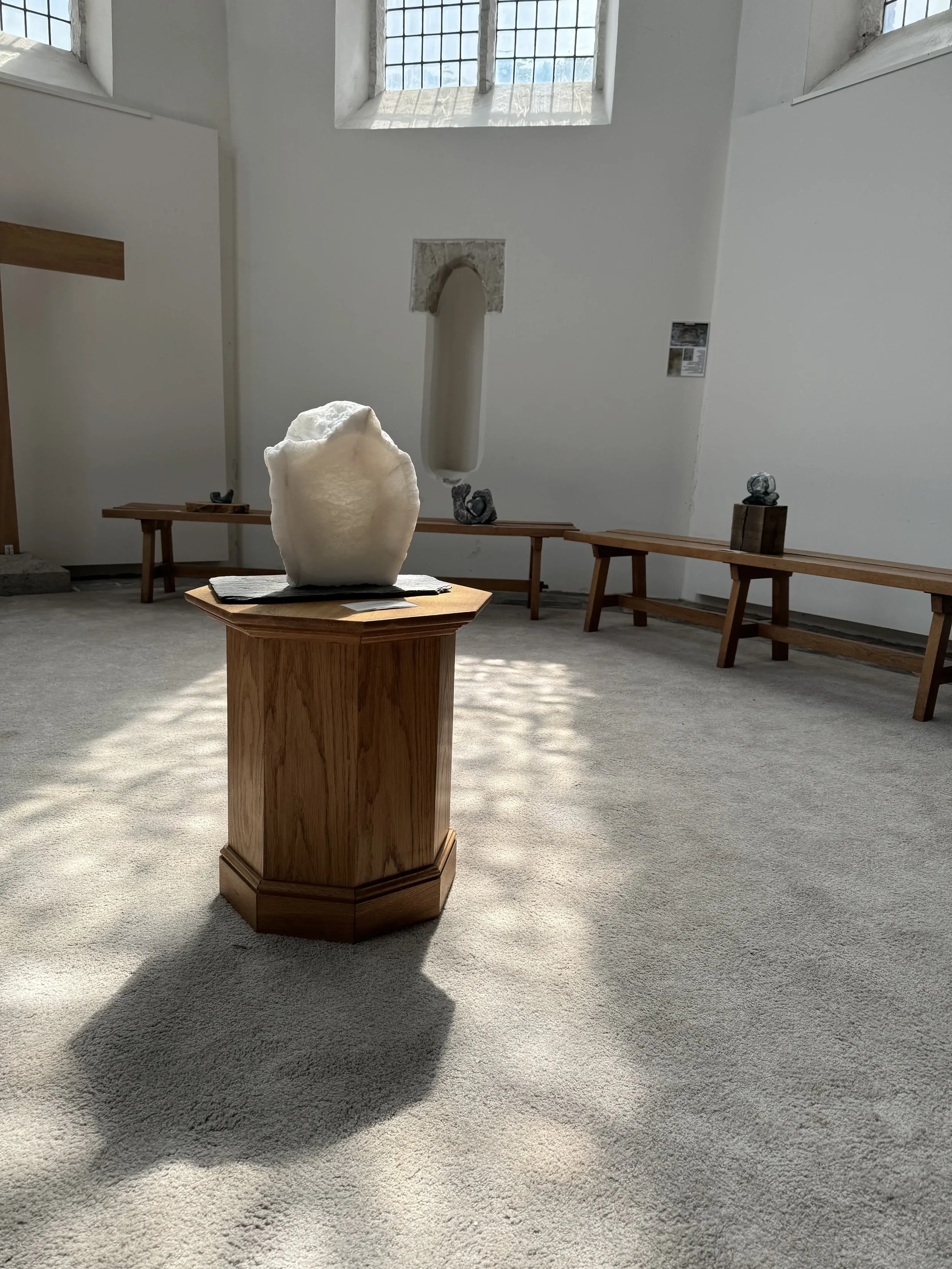 Een galerij met sculpturen in een ruimte met grote ramen. Op een houten standaard staat een witte, ruwe sculptuur. Op de achtergrond staan andere kleine sculpturen op houten banken.