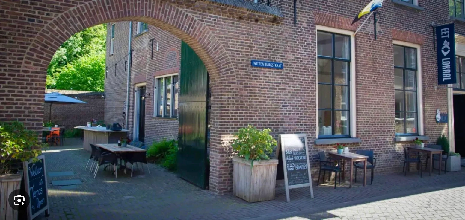 Uitzicht op een gezellige terras met buitentafels en stoelen voor een restaurant, gelegen in een historisch gebouw met een stenen boog en ramen met planten en menu's. Een bord met de naam Wittenburgstraat is zichtbaar.