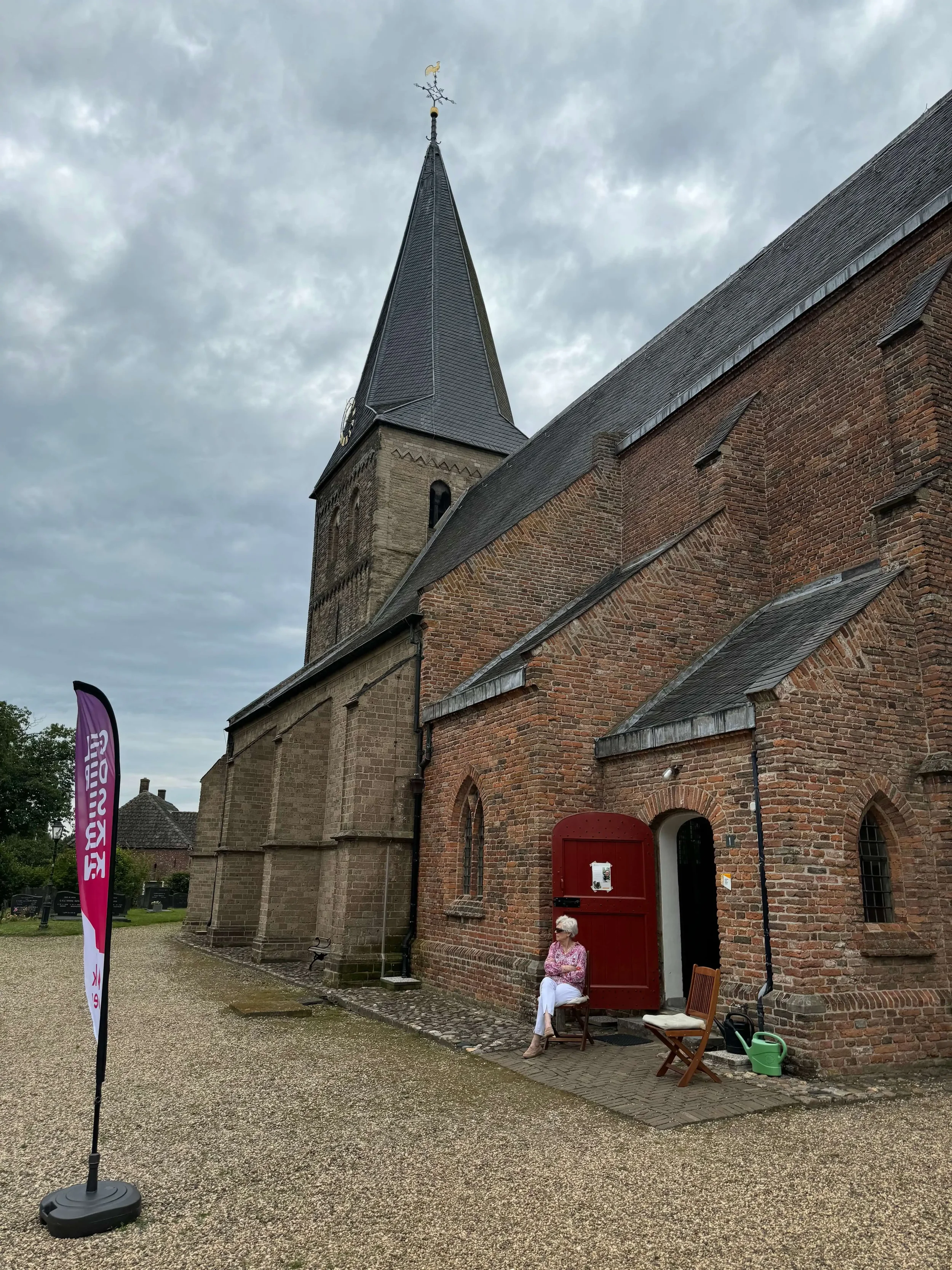 Een oude kerk met een hoge toren en een regenachtige lucht op de achtergrond. Voor de kerk zit een vrouw op een stoel, met naast haar een aantal stoelen en een tuiniergereedschap, een felrood deur en een vlaggenmast.