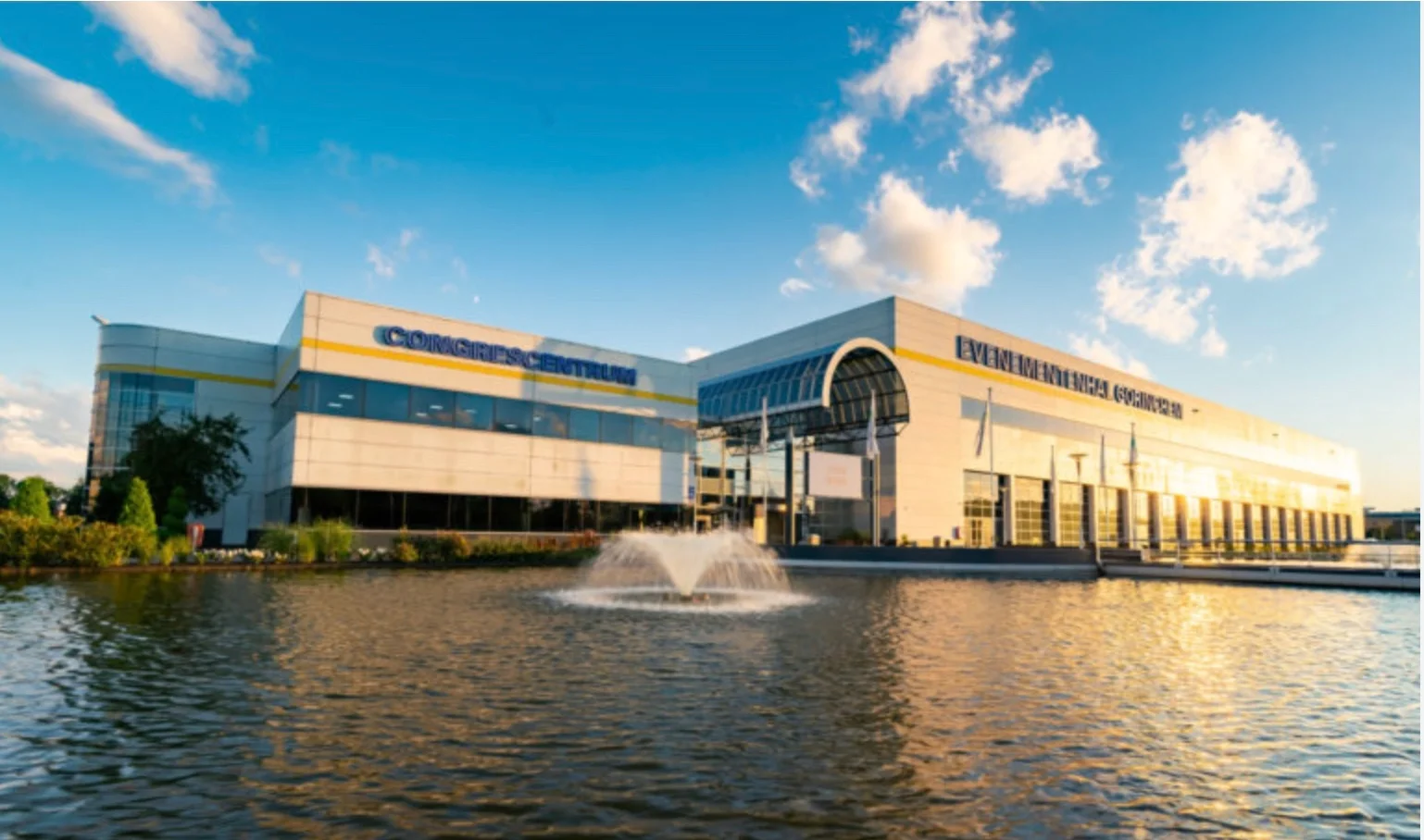 Modern evenementencentrum met een waterpartij en fontein, onder een blauwe hemel met wolken.