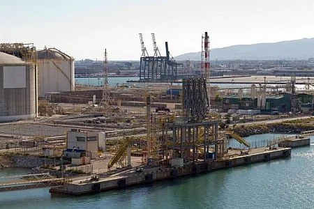 Industrial port with cranes, pipelines, and storage tanks near water.