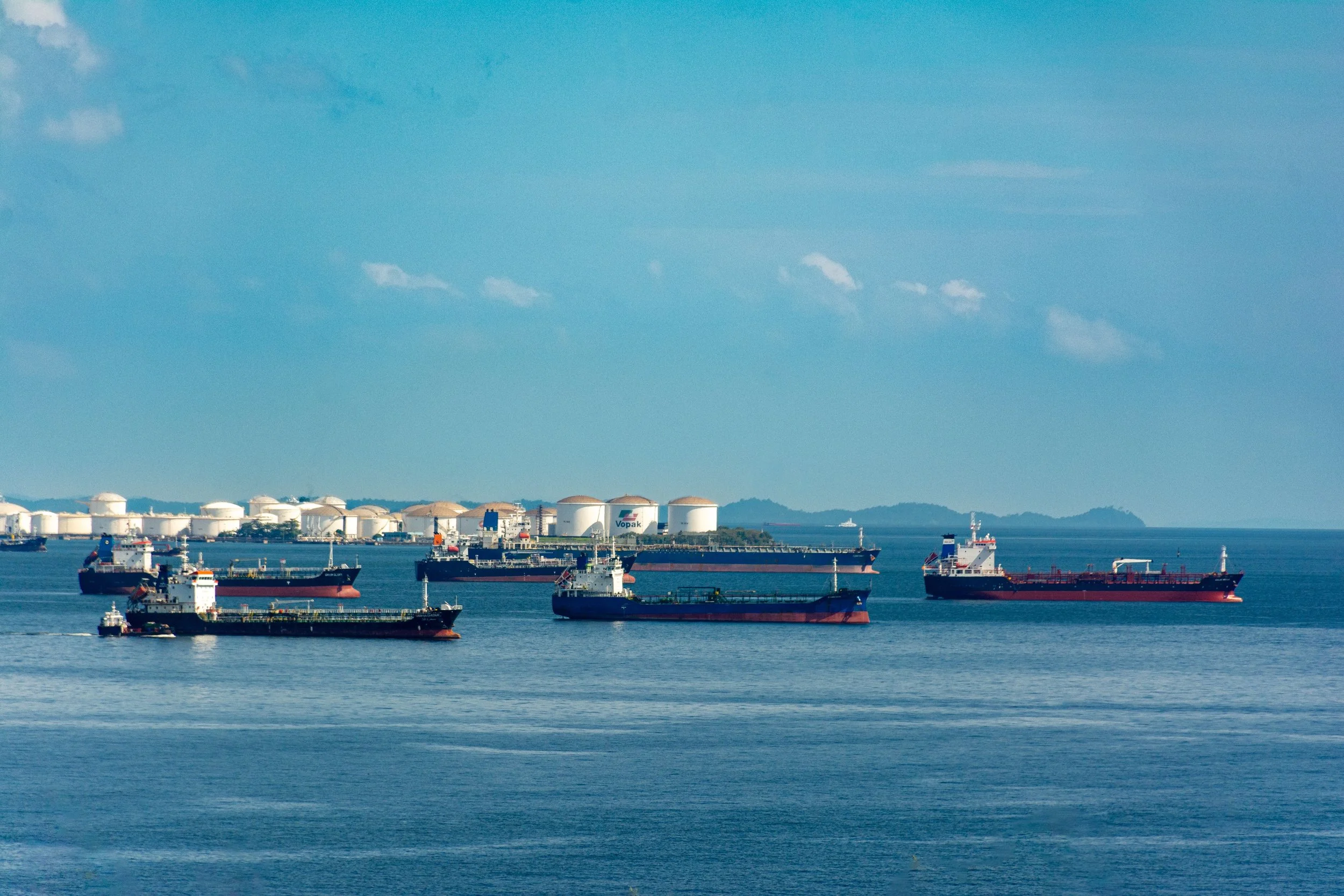A harbor with multiple large cargo ships and tankers floating on calm water, with oil storage tanks and a distant shoreline under a partly cloudy sky.