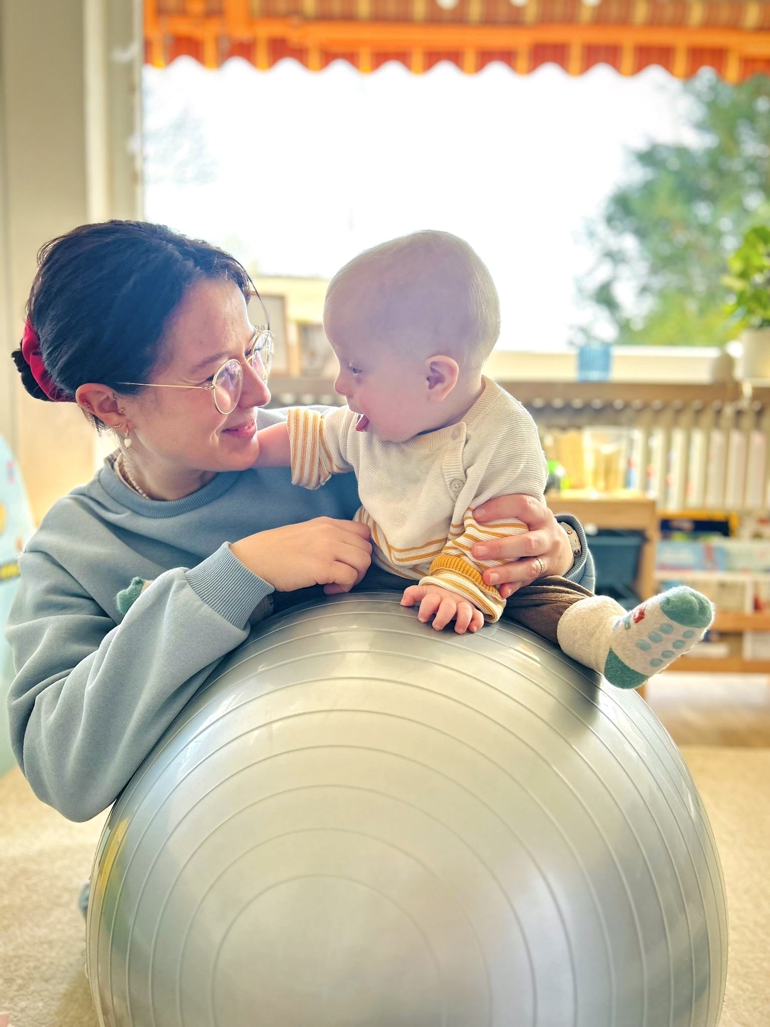 Eine Frau hält einen kleinen Jungen auf einem Gymnastikball in einem Raum mit Bibliothek im Hintergrund. Die Frau trägt Brille und hat dunkle Haare, der Junge trägt eine gelbe Pumphose und bunte Socken.