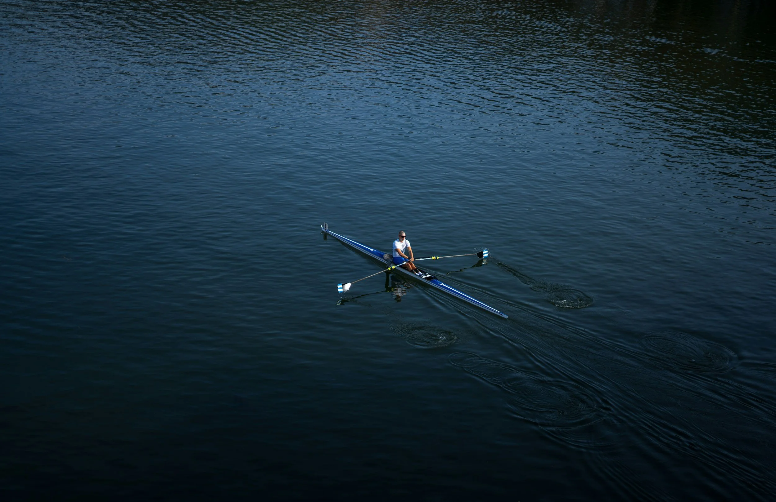 A person wearing a white shirt and a cap rowing a narrow racing shell on a calm lake.