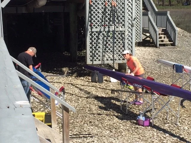 Two people preparing a purple shell on a metal stand, with a third person nearby, under a wooden deck with stairs, on a pebbled ground