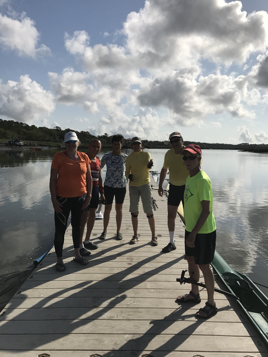 Group of six people standing on a wooden dock next to a body of water, with boats and rowing shells nearby, under partly cloudy sky.