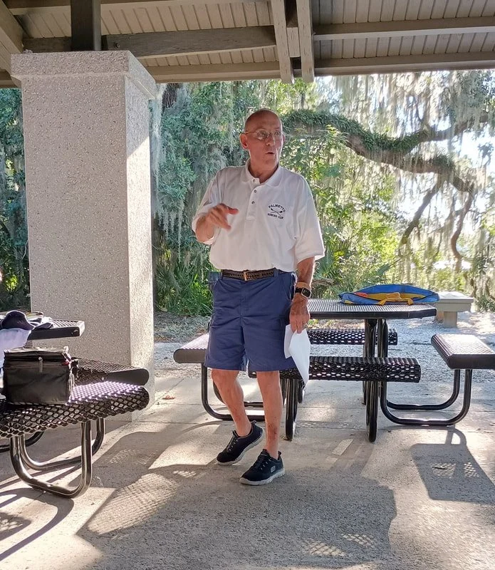 A man dressed in a white polo shirt and blue shorts standing under a pavilion, holding papers and gesturing with his right hand, with a background of trees and hanging moss.