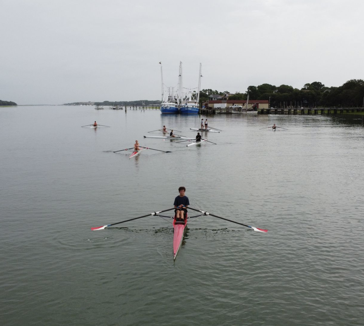 A boy rowing a single scull for Palmetto Rowing Club on Broad Creek, Hilton Head Island, with other rowers in the background and boats docked by the shoreline.