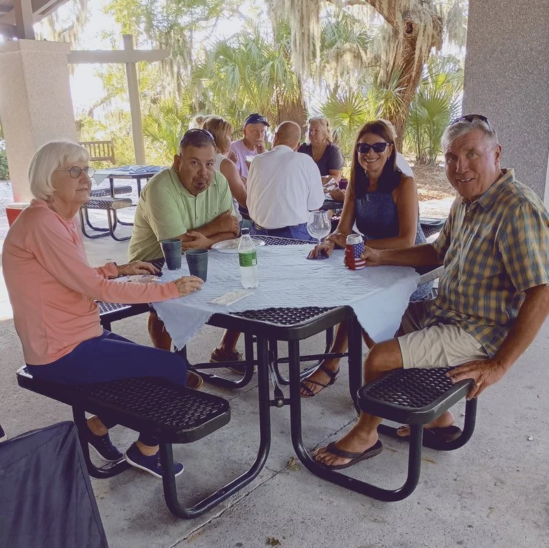 Group of eight members of the Palmetto Rowing Club sitting around a picnic table outdoors, with trees and foliage in the background, enjoying a social gathering.