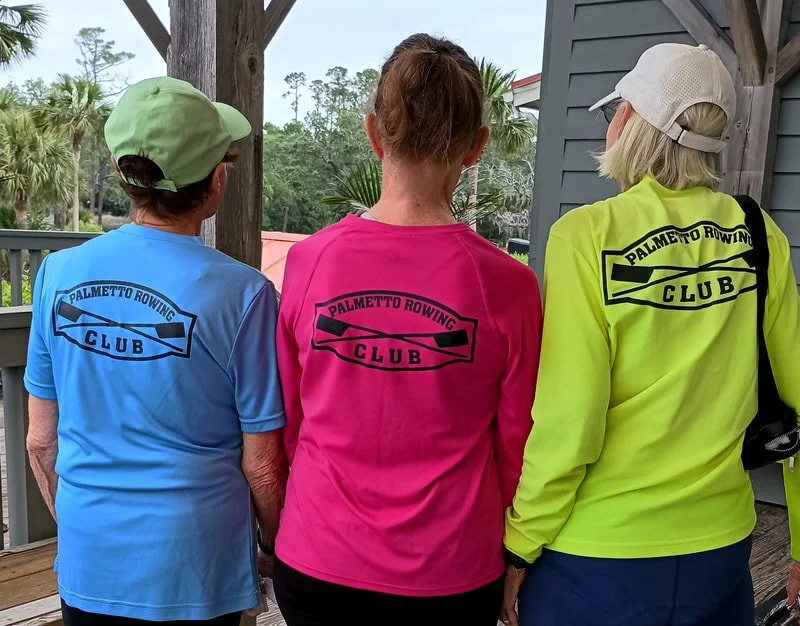 Three women wearing colorful shirts for Palmetto Rowing Club and hats, standing outdoors on a wooden deck, with trees and a building in the background.