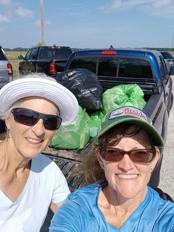 Two women taking a selfie in front of a pickup truck loaded with black and green garbage bags in an open outdoor area under a clear sky.