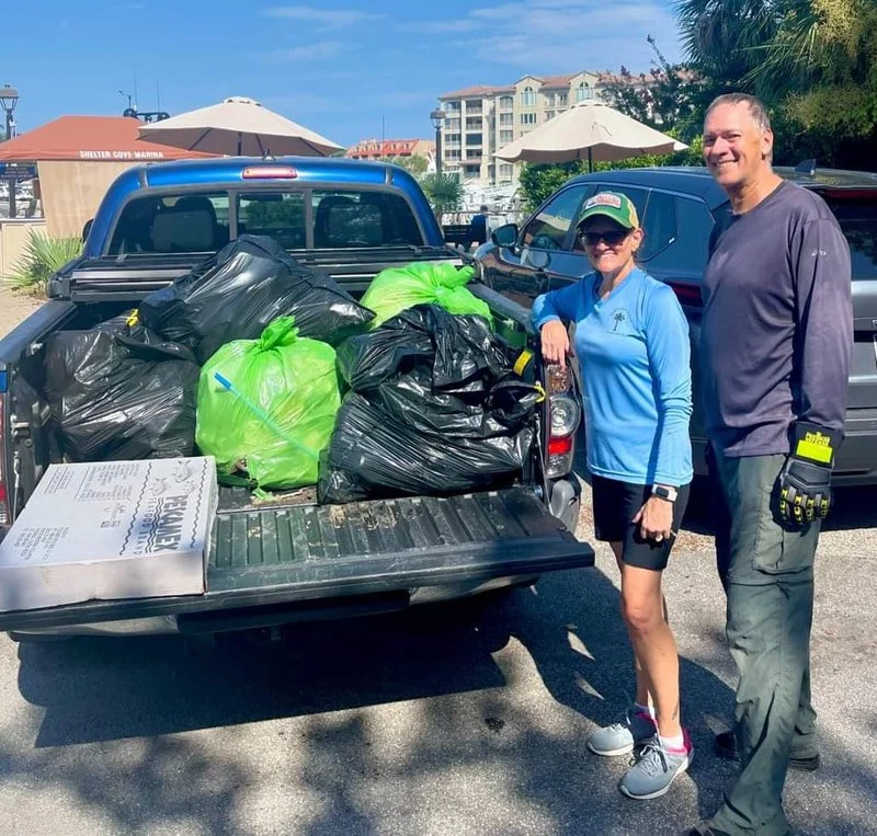 Two people, a woman and a man, stand next to a pickup truck filled with black and green trash bags. They are smiling, wearing outdoor work clothing, and appear to have participated in a cleanup event. The background shows a parking lot with umbrellas