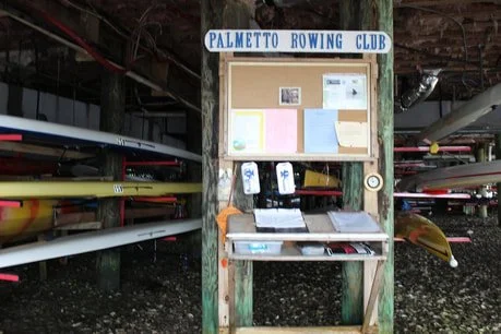 Bulletin board and storage area for Palmetto Rowing Club with rowing shells and paddles nearby.