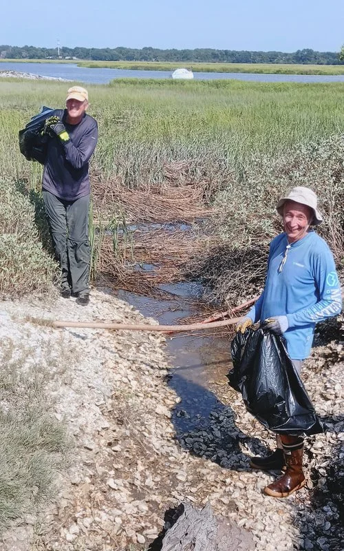 Two women standing in a marshy area near a water body, engaging in a cleanup activity. One woman is wearing a hat, gloves, and holding a large black trash bag, while the other is dressed in outdoor clothing and gloves, carrying a black bag and a back
