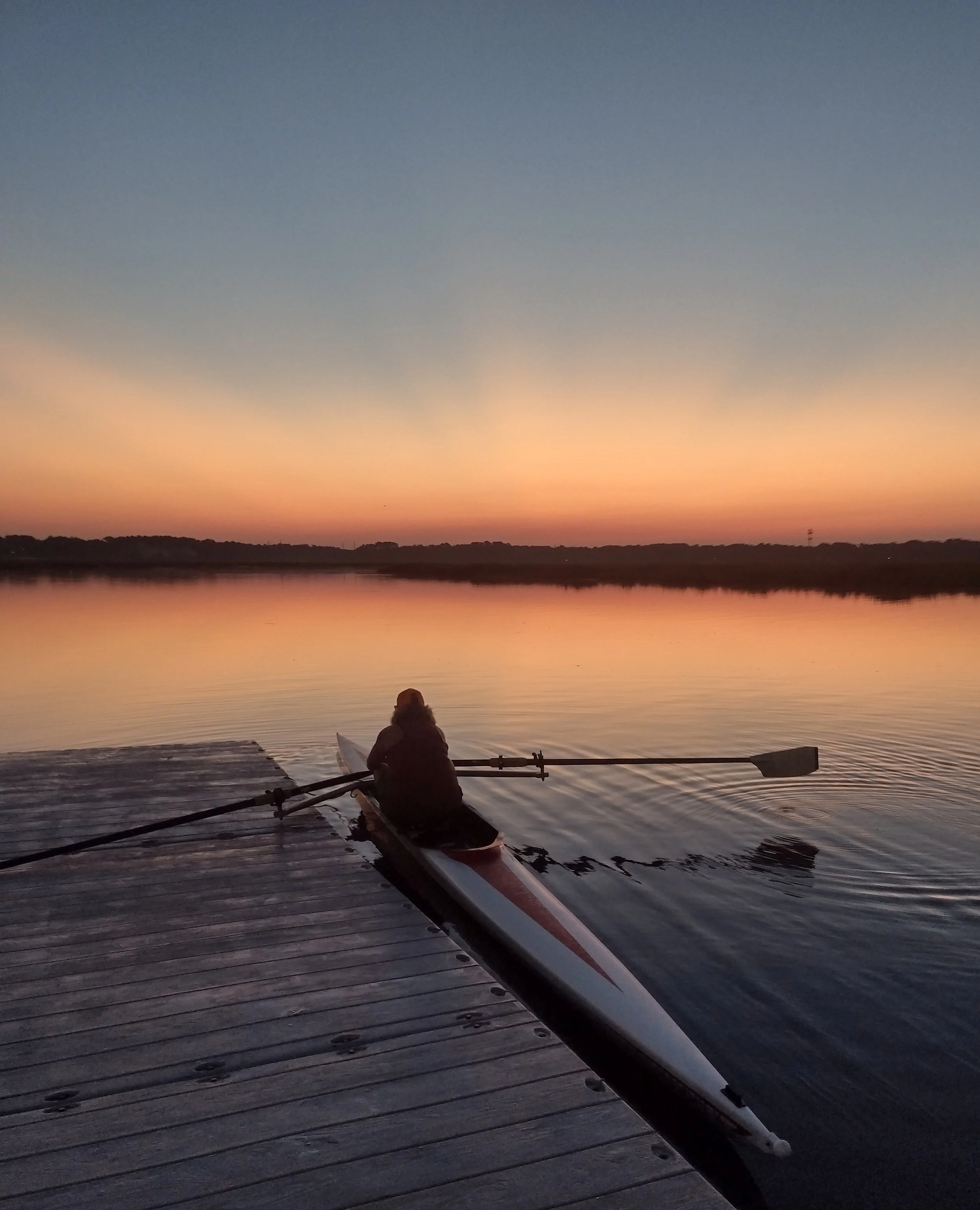 Person in a boat on a calm river at sunset, with a colorful sky and distant trees and structures.