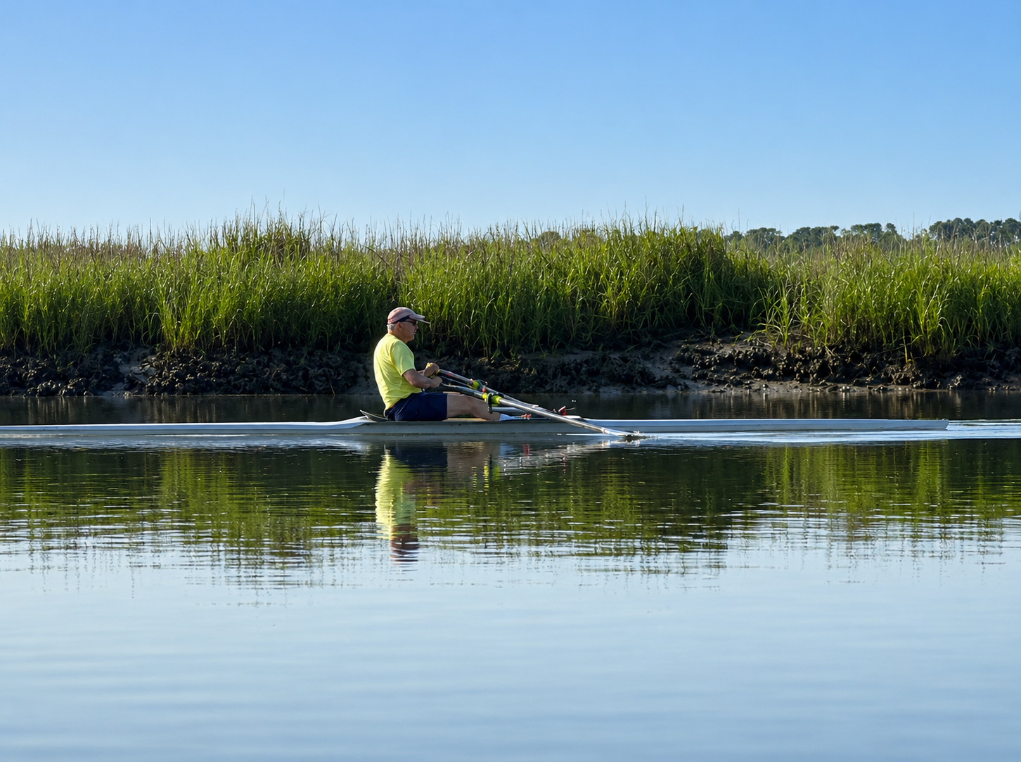 A person in a yellow shirt and shorts rowing a narrow boat on calm water, with tall green grass and trees in the background under a clear blue sky.