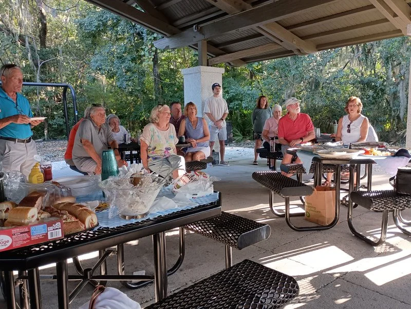 Group of people gathered under a pavilion for a social outdoor meal, with food and drinks on tables, surrounded by trees.