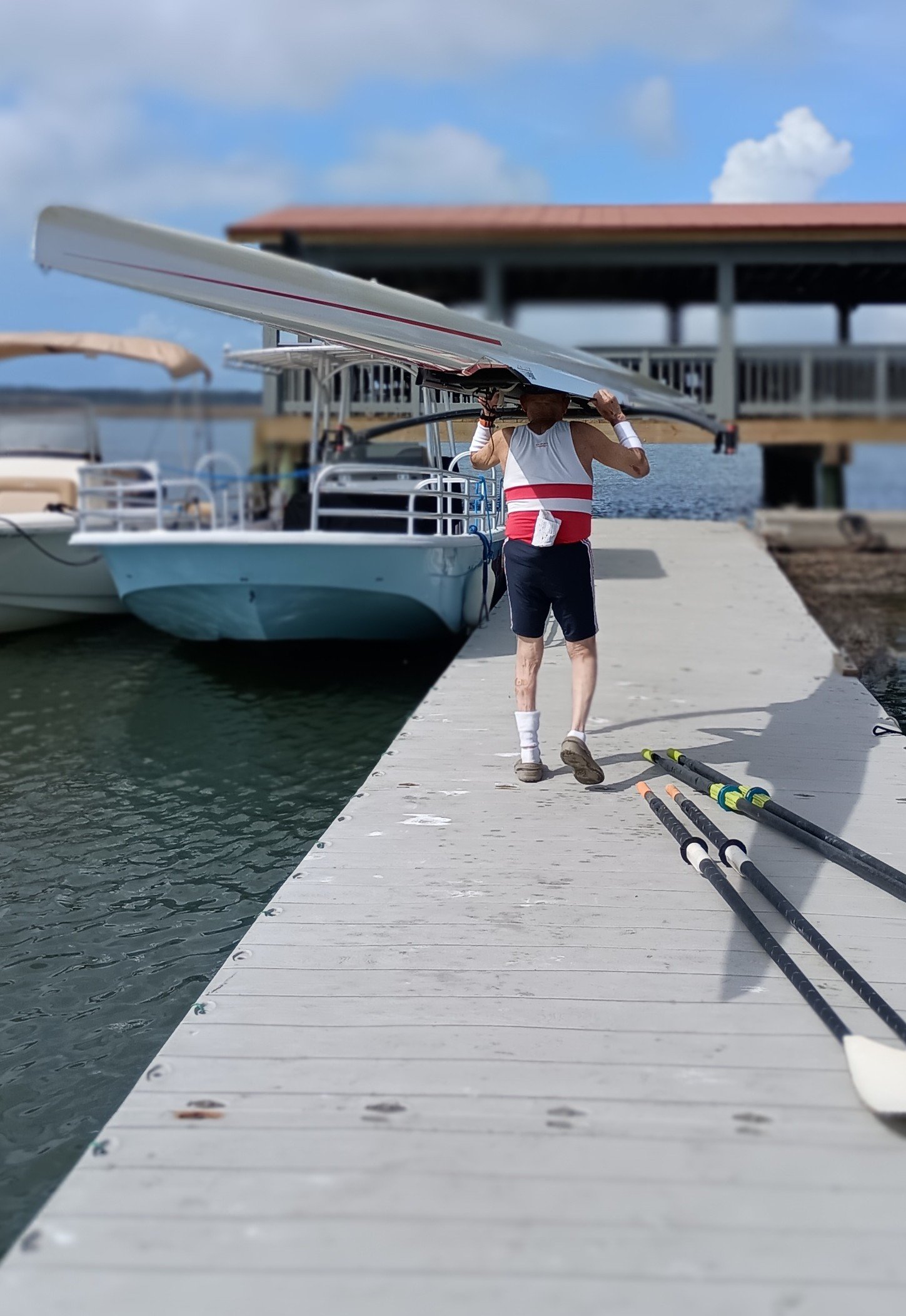 A person dressed in athletic clothing carrying a shell on their shoulders along a docked pier by the water, with paddles lying on the ground nearby.