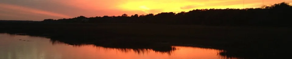 Sunset over a calm river with a silhouette of trees in the distance.
