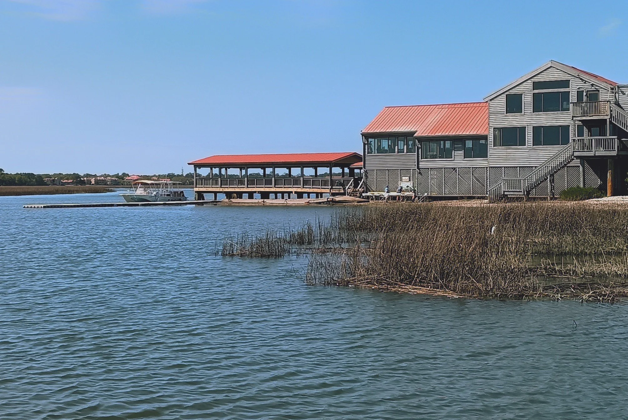 A waterfront scene with a house on stilts, a porch extending over the water, and a boat docked nearby on a sunny day.