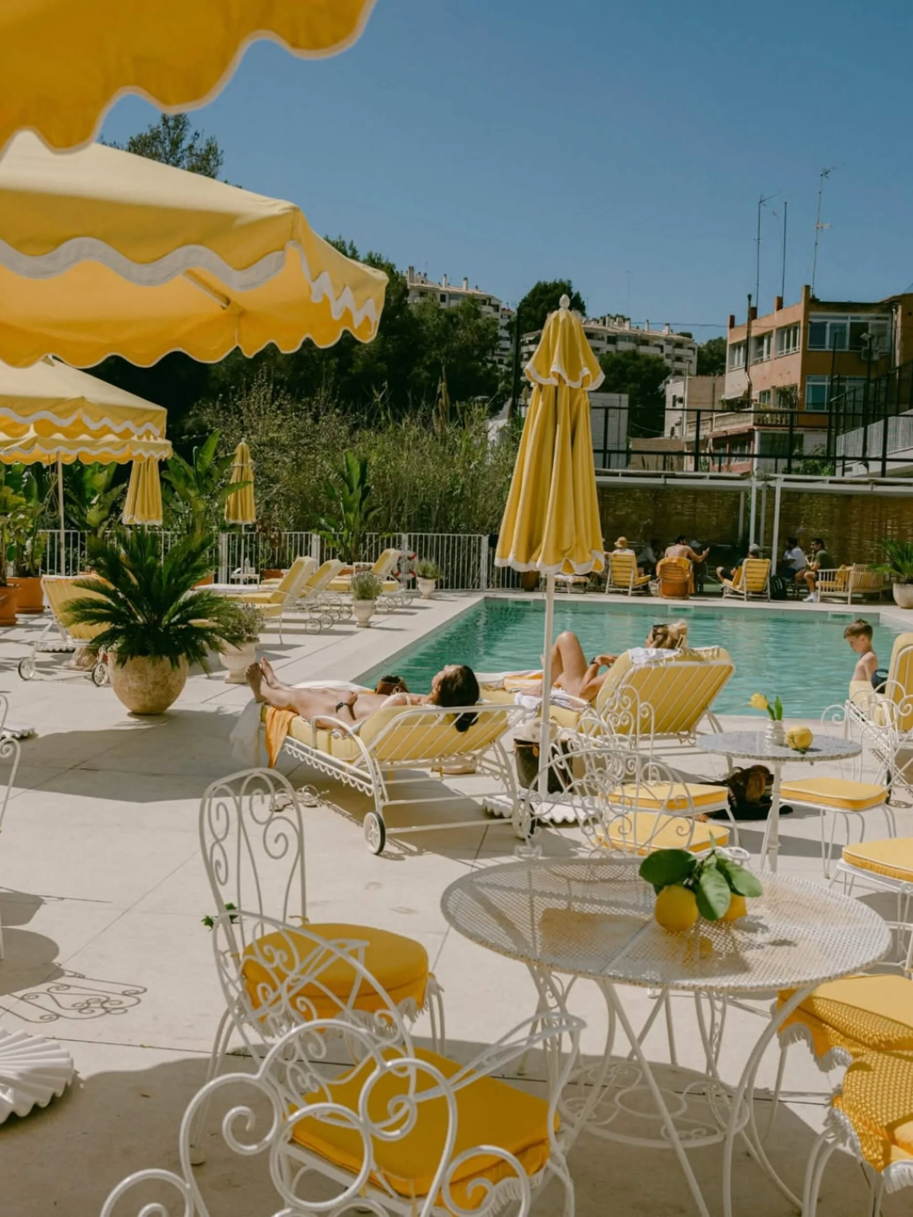 People relaxing and sunbathing by a swimming pool with yellow and white patio furniture, umbrellas, and green plants, in an outdoor setting with buildings in the background on a sunny day.