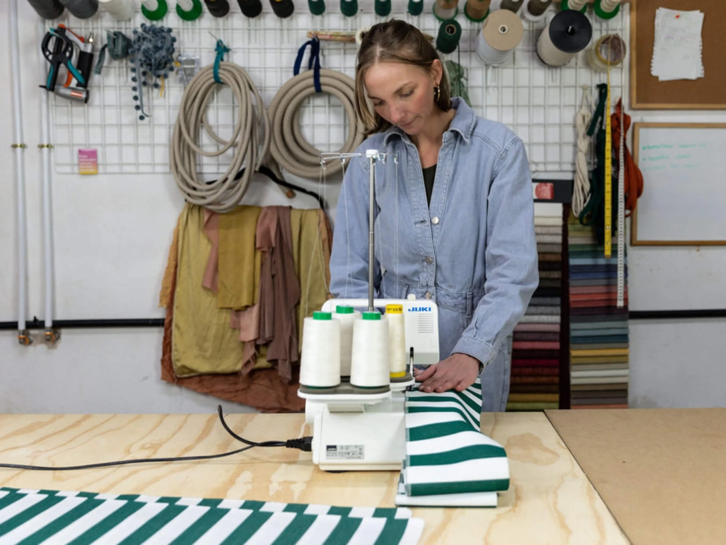 A woman in a denim shirt working with green and white striped fabric on a sewing machine in a craft room.
