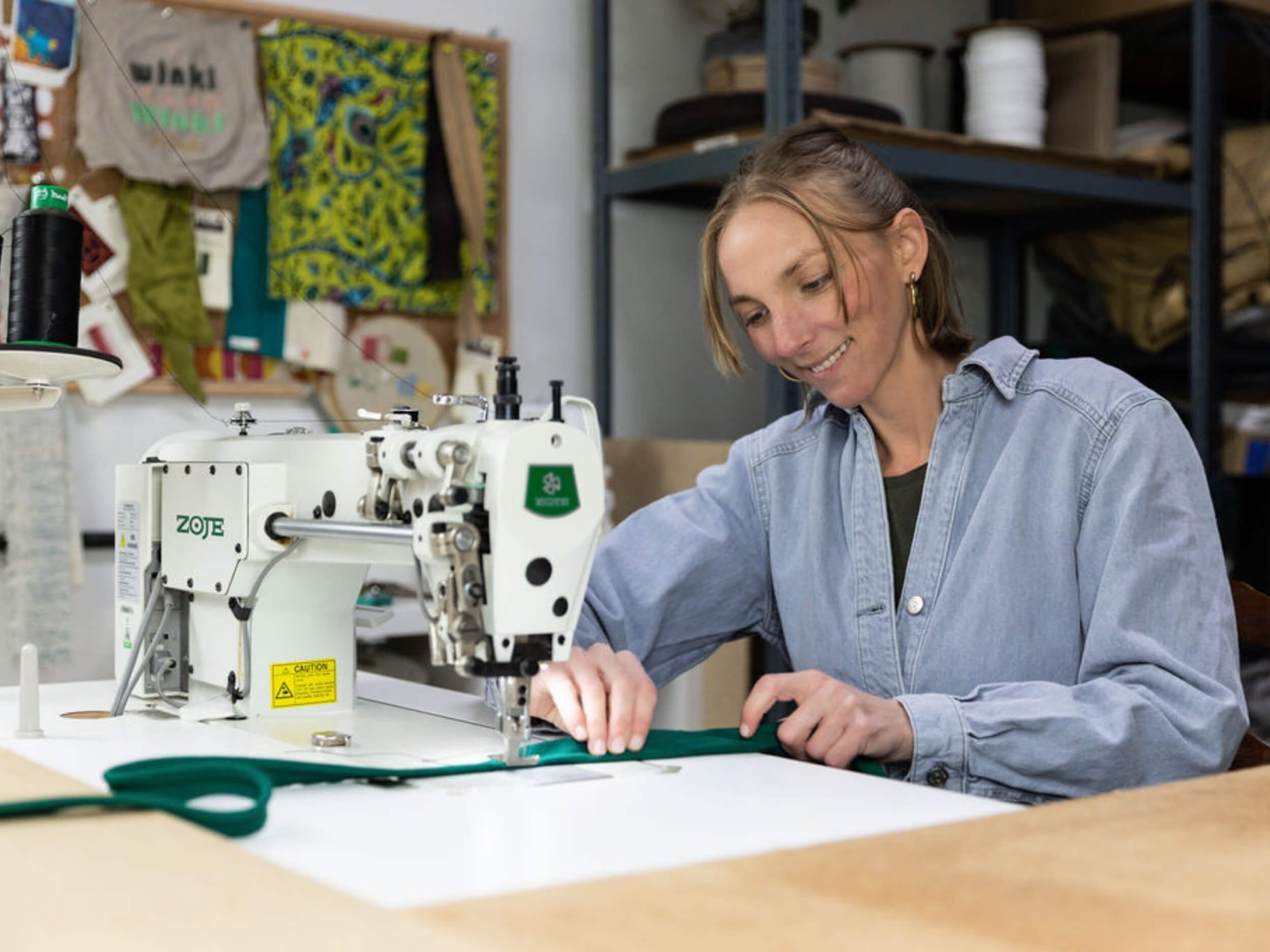 A woman working with a sewing machine in a workshop, surrounded by fabric and sewing supplies.
