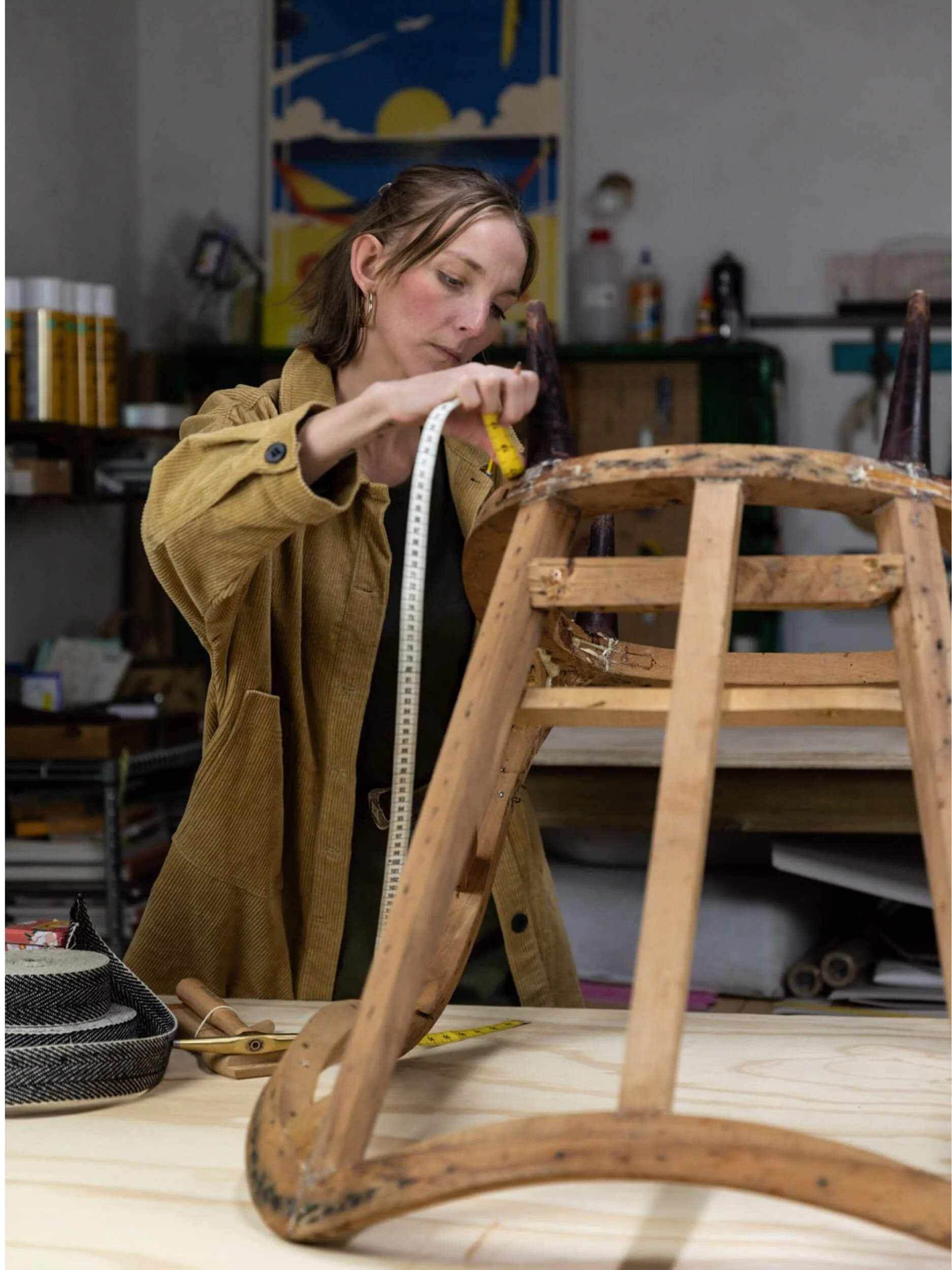Woman working on restoring a wooden chair in a workshop, with tools and supplies on the table and shelves in the background.