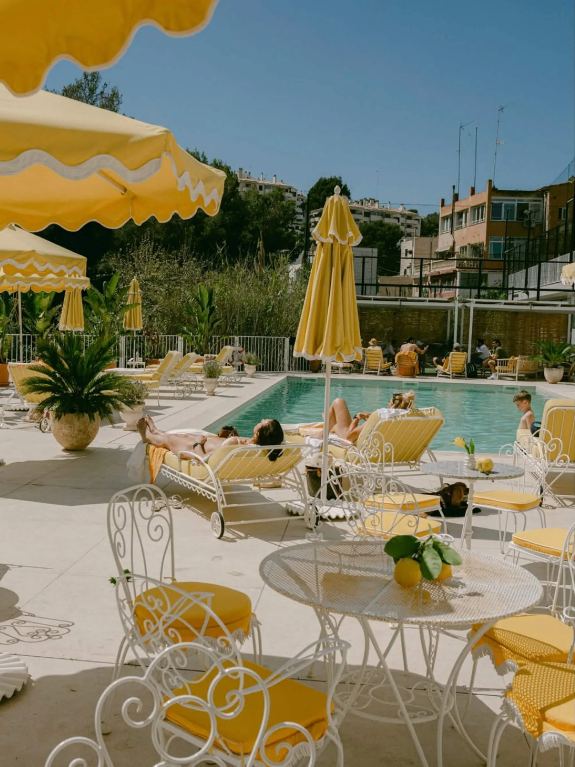 Outdoor poolside area with yellow umbrellas and lounge chairs, people relaxing, surrounded by plants and buildings in the background.
