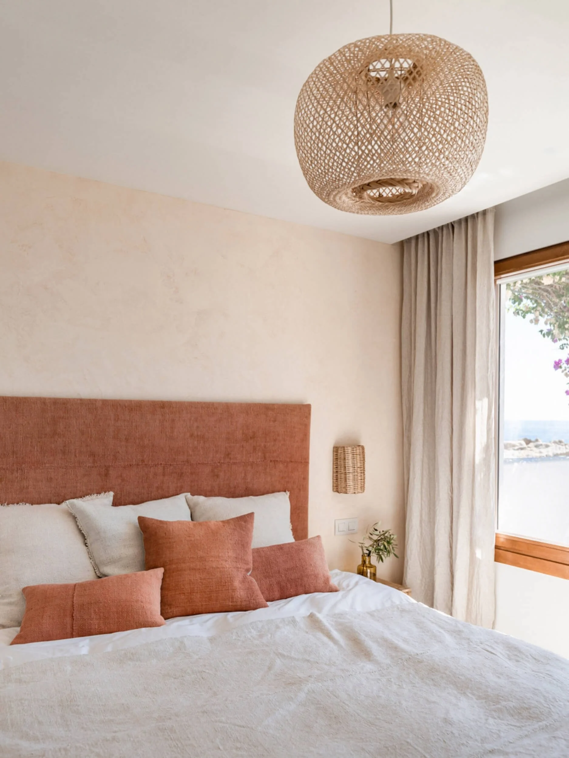 A bedroom with a bed, six peach-colored pillows, a beige headboard, a small side table with a plant, a wicker wall sconce, light-colored curtains, and a view outside of trees and the ocean.