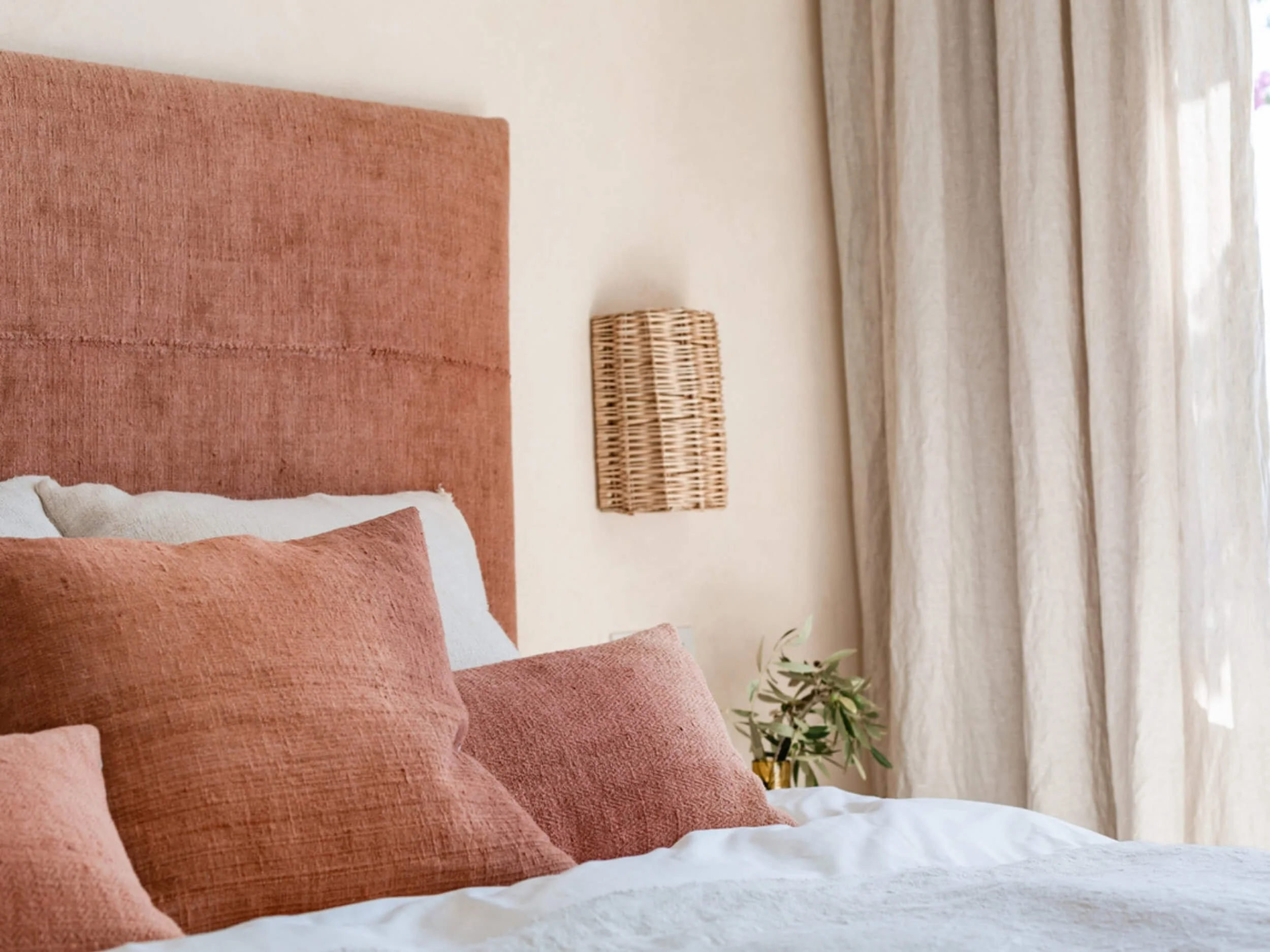 A bedroom with a pink upholstered headboard, matching pink pillows, a wicker wall decoration, a small nightstand with a gold vase holding greenery, beige curtains, and soft natural lighting.