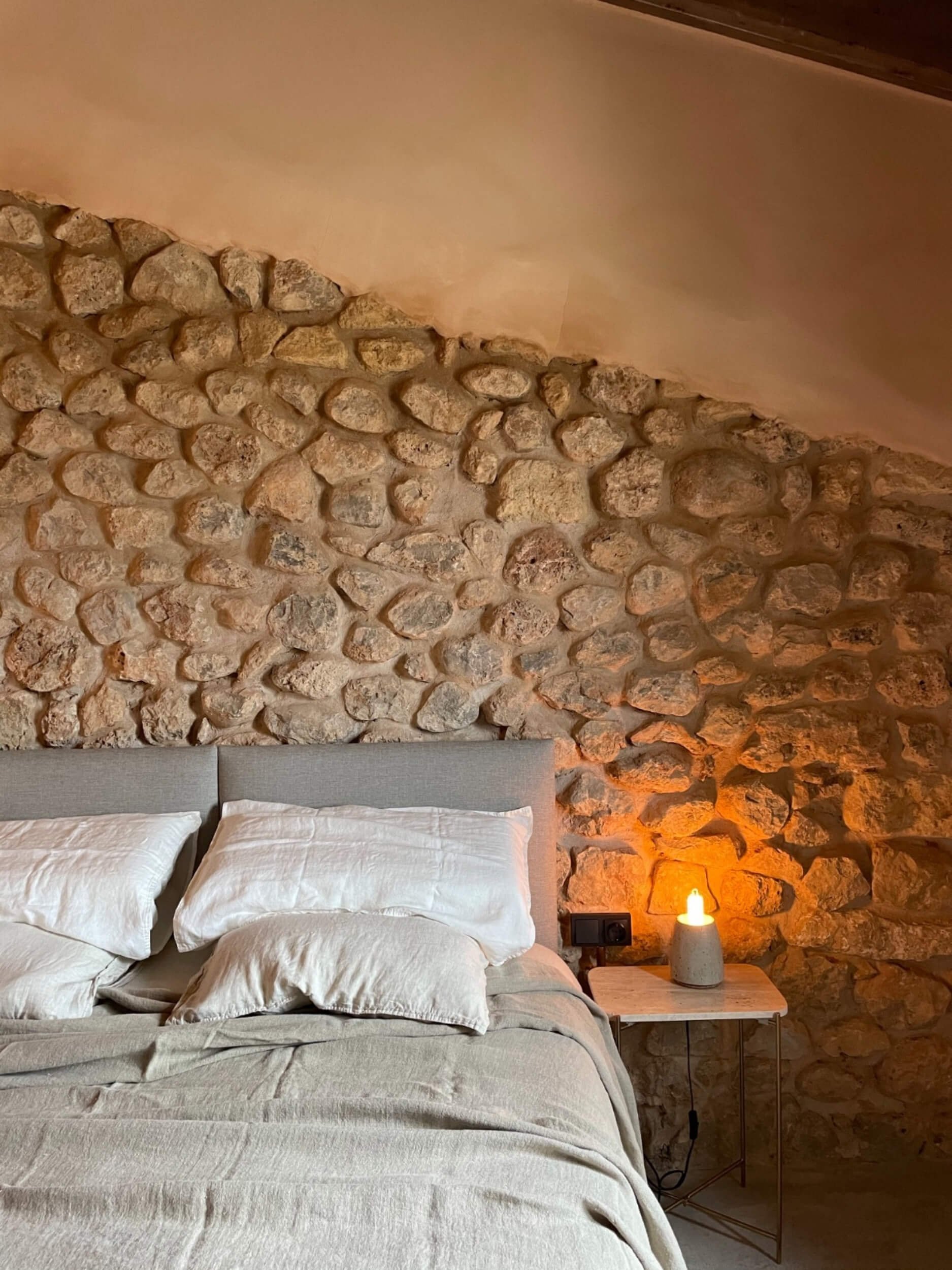 A cozy bedroom with a stone accent wall behind a bed with white bedding and pillows, a wooden side table with a lamp, and warm ambient lighting.