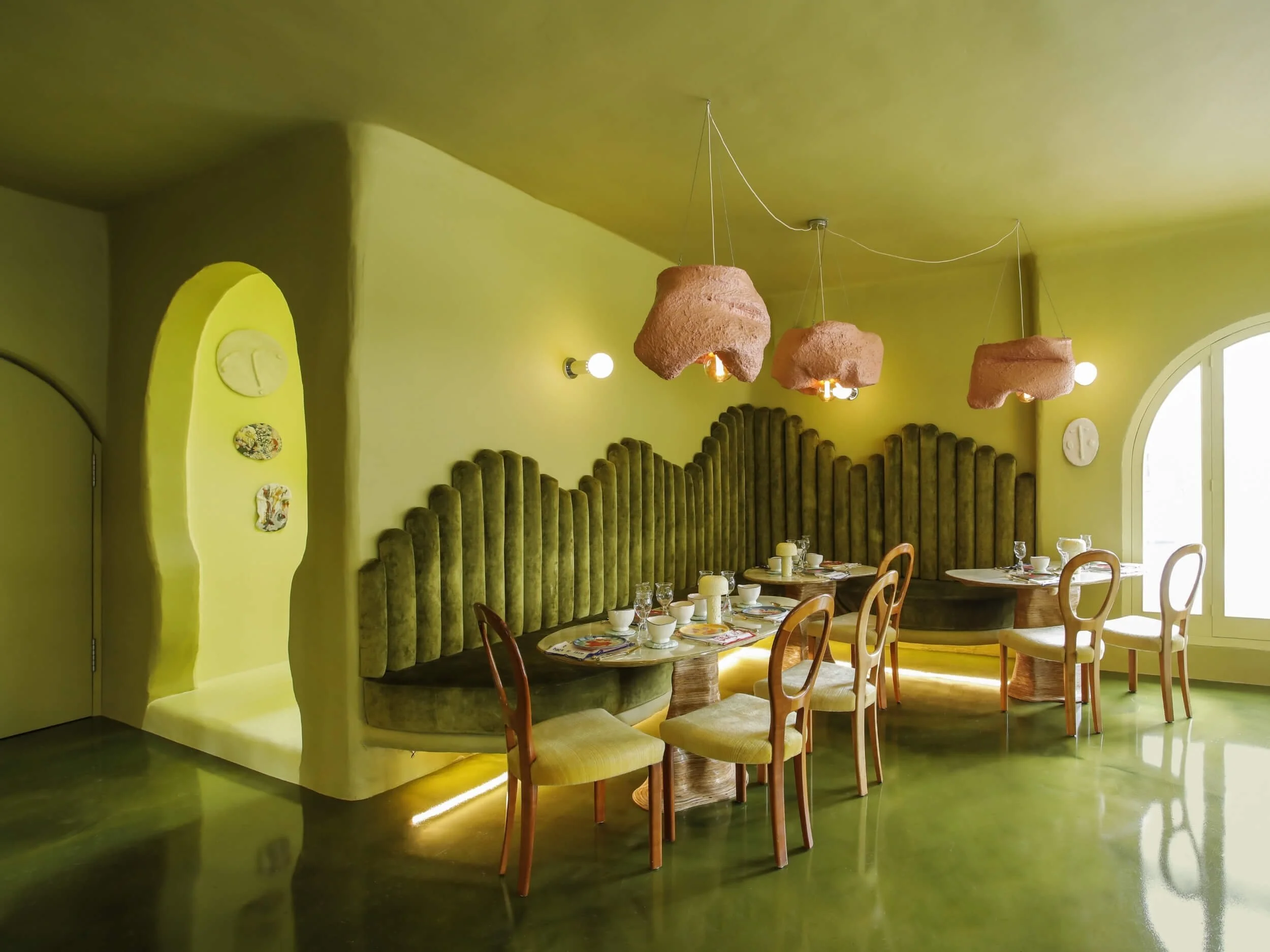 A brightly lit dining area with yellow-green walls and a matching floor. There are wooden chairs surrounding a long table set with white cups, plates, and utensils. Hanging from the ceiling are three unique pinkish-brown pendant lights. The room features a curved wall with attached decorative plates and an arched window allowing natural light.