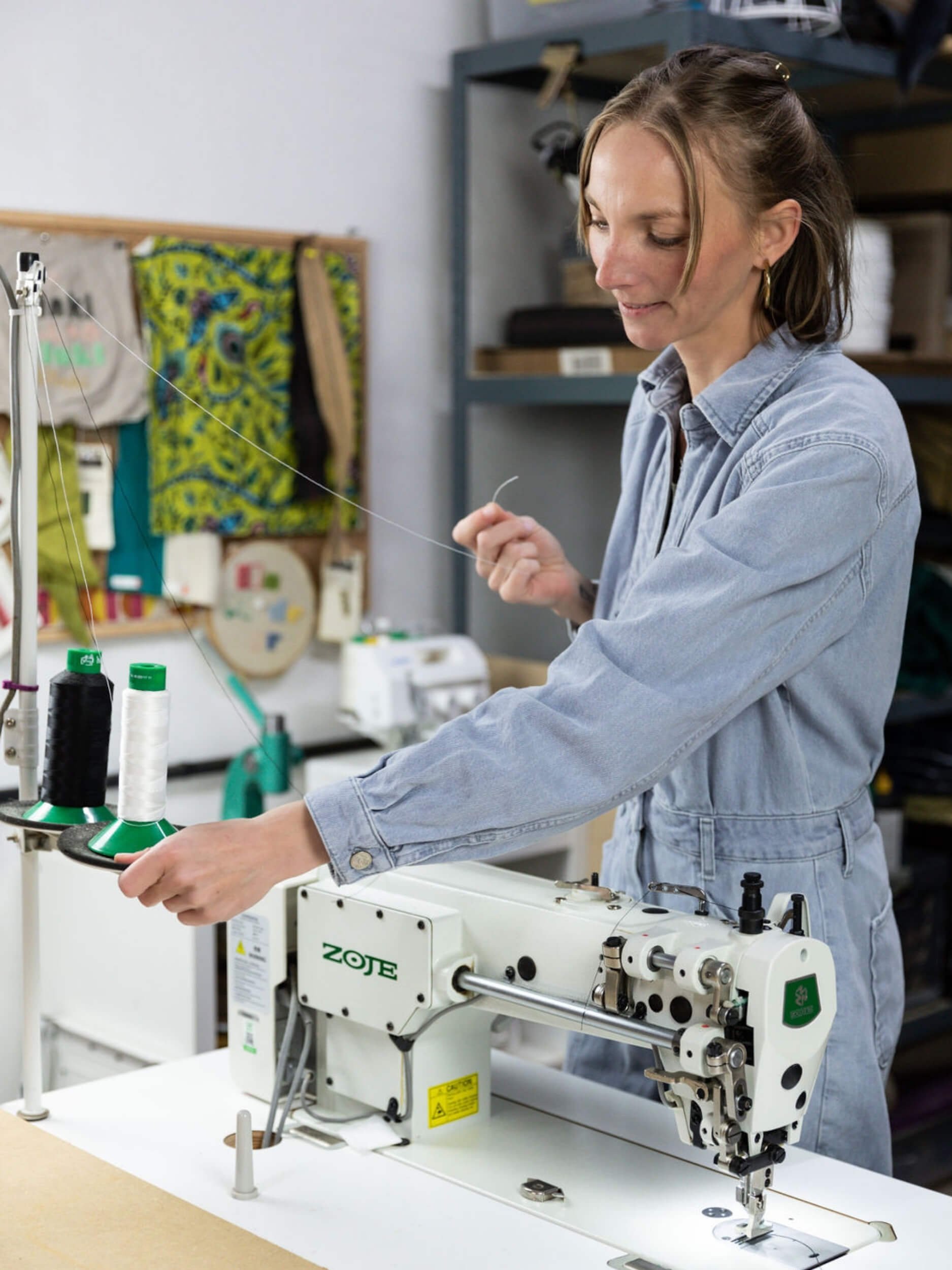 A woman in a light blue denim shirt operates a textile sewing machine in a workshop, with sewing thread spools in the foreground and shelves in the background.