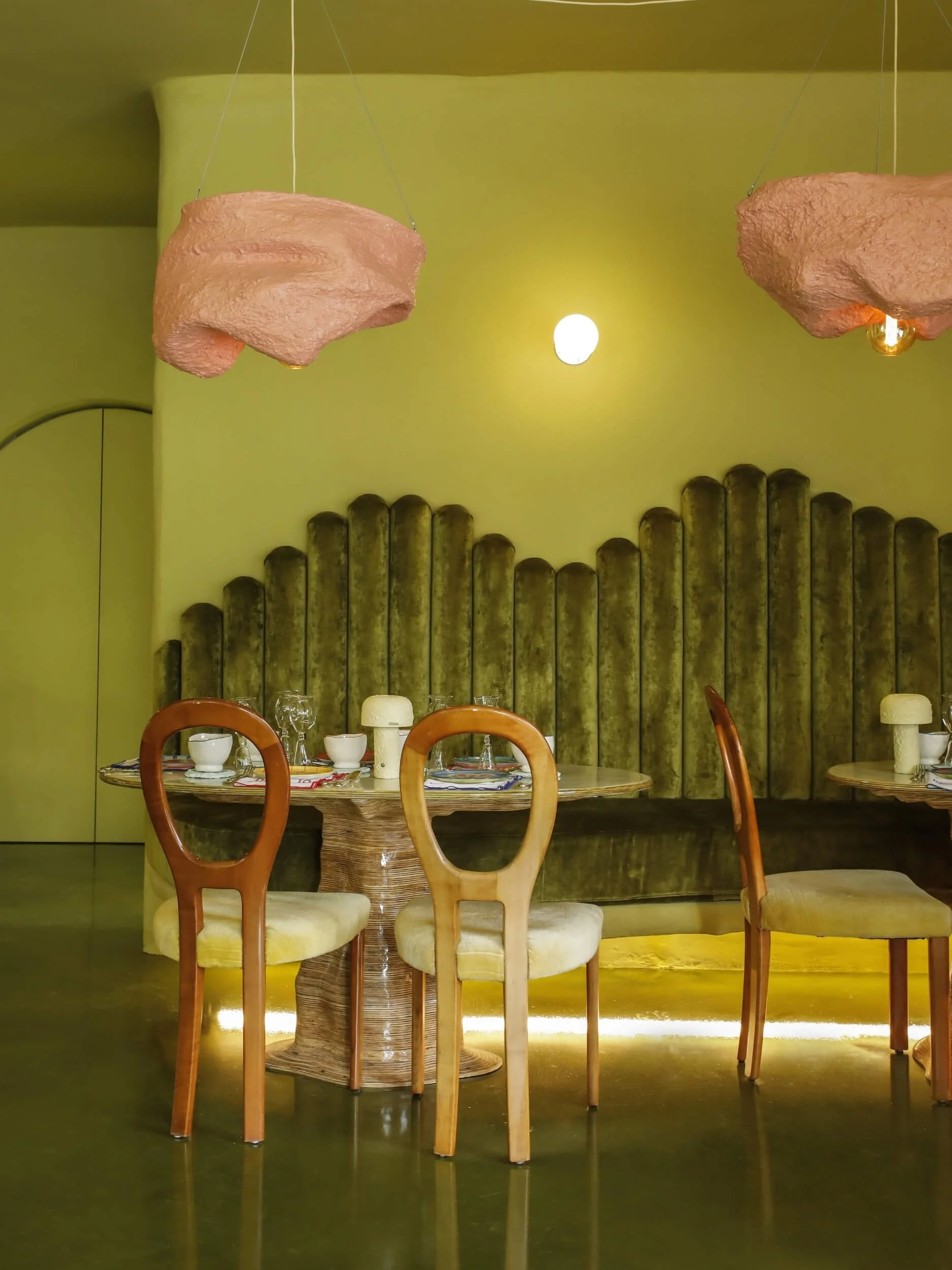 A set of four wooden chairs with cream-colored cushions surrounds a round table set for dining, with white bowls and glasses, in a restaurant with a green wall and decorative hanging lights shaped like pink rocks with embedded light bulbs.