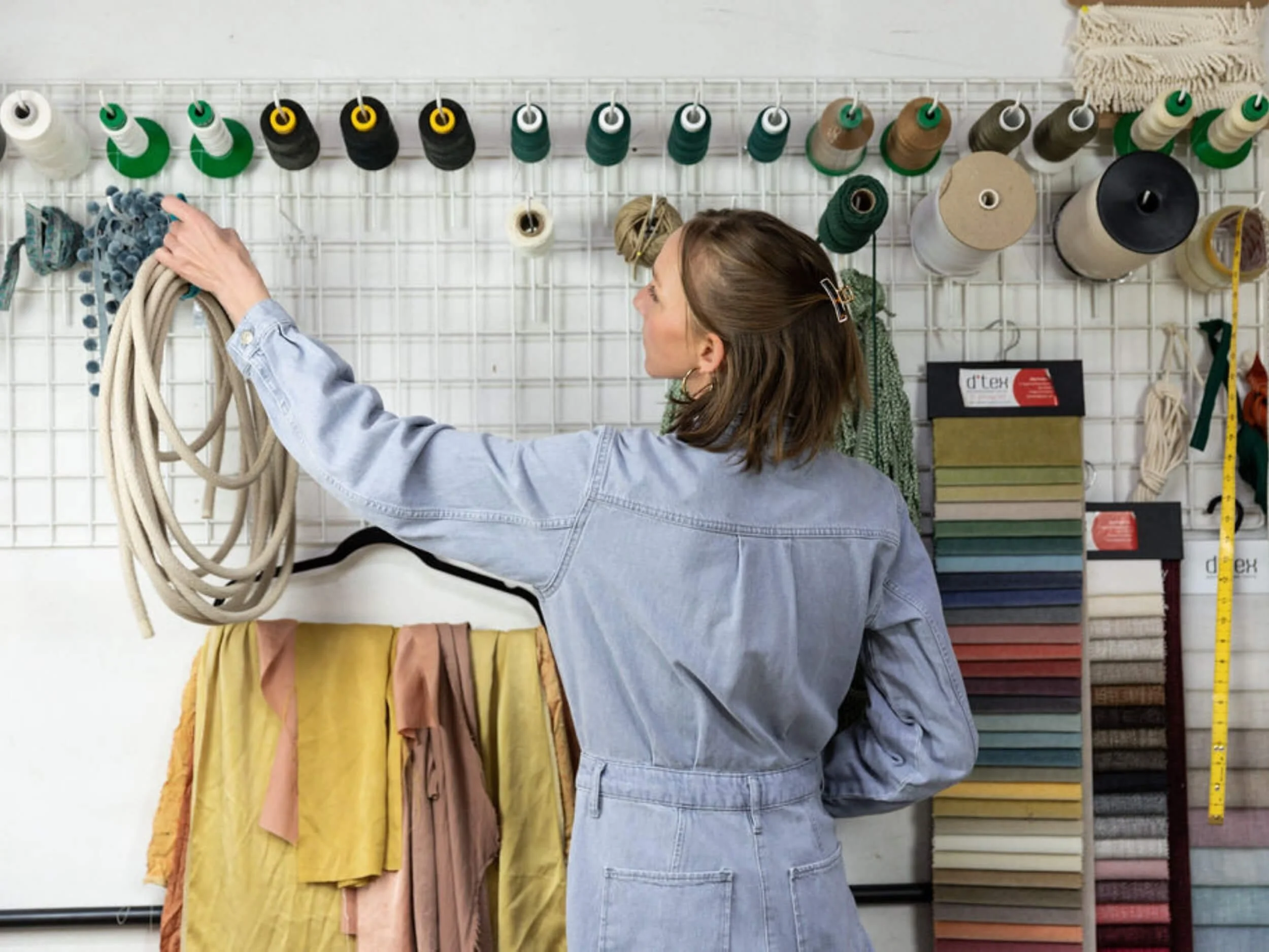 A woman with brown hair in a denim jacket handles colorful fabric swatches in a craft or fabric store, with spools of thread and rolls of fabric on a white grid wall and a yellow measuring tape in the background.