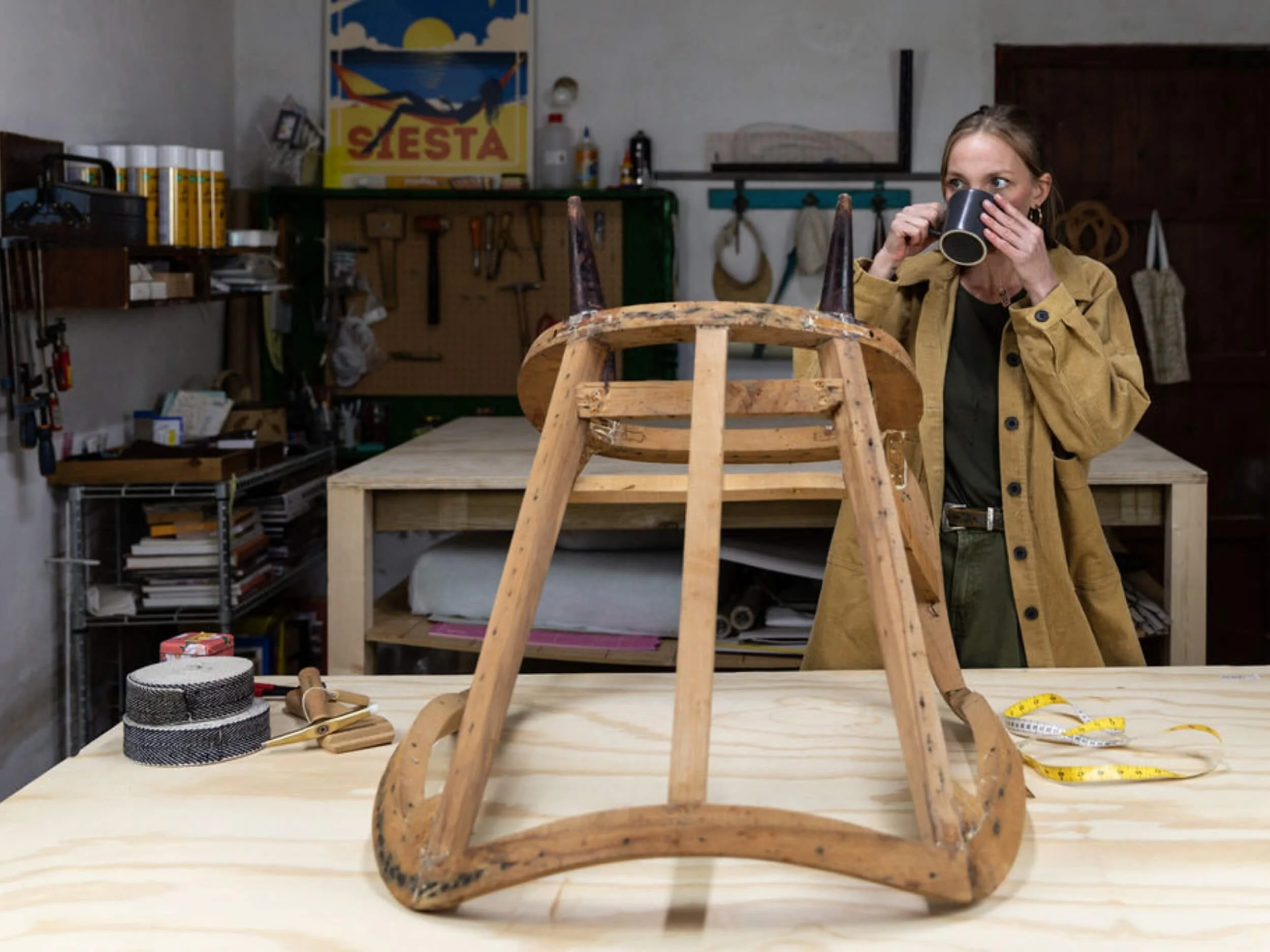 A woman in a workshop holding a black cup, observing a small wooden chair frame on a table.