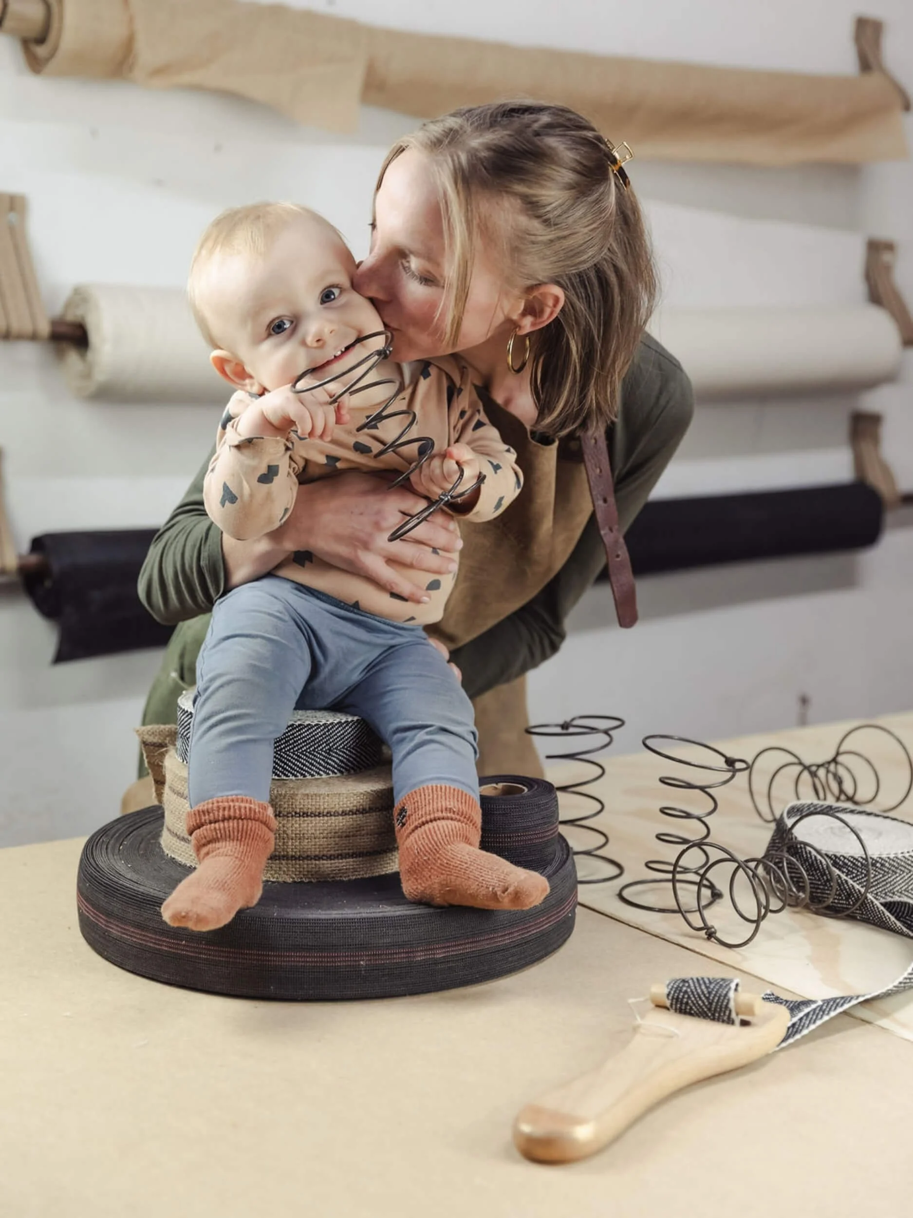 A woman is hugging and kissing a young boy, holding him close with her eyes closed. The boy is smiling, holding a pair of glasses. They are standing behind a table with multiple rolls of black and gray ribbon, a spool of black wire, and other crafting supplies, with a wooden background behind them.