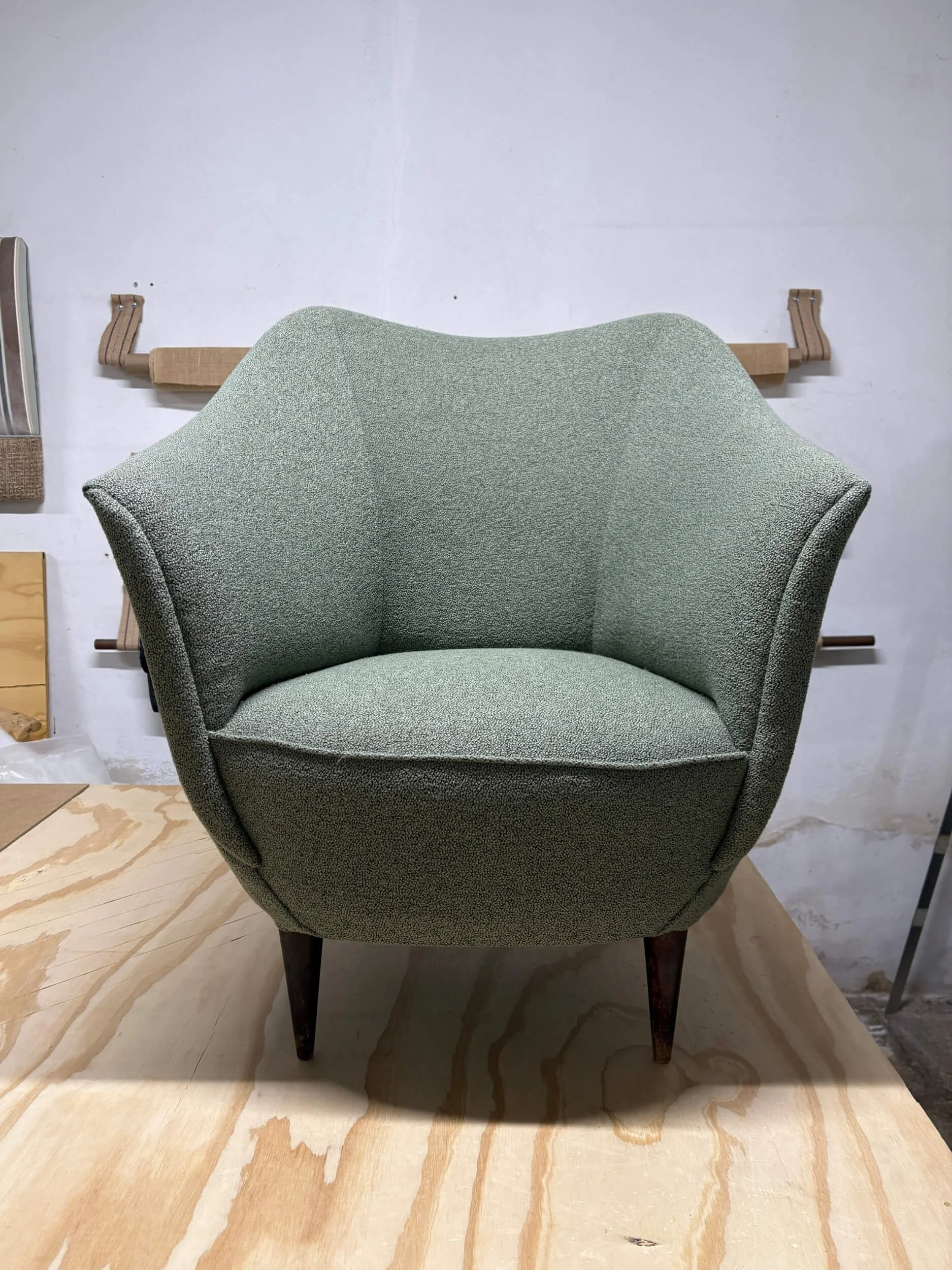 An upholstered armchair with dark wooden legs in front of a plain white wall, on a light wooden floor.