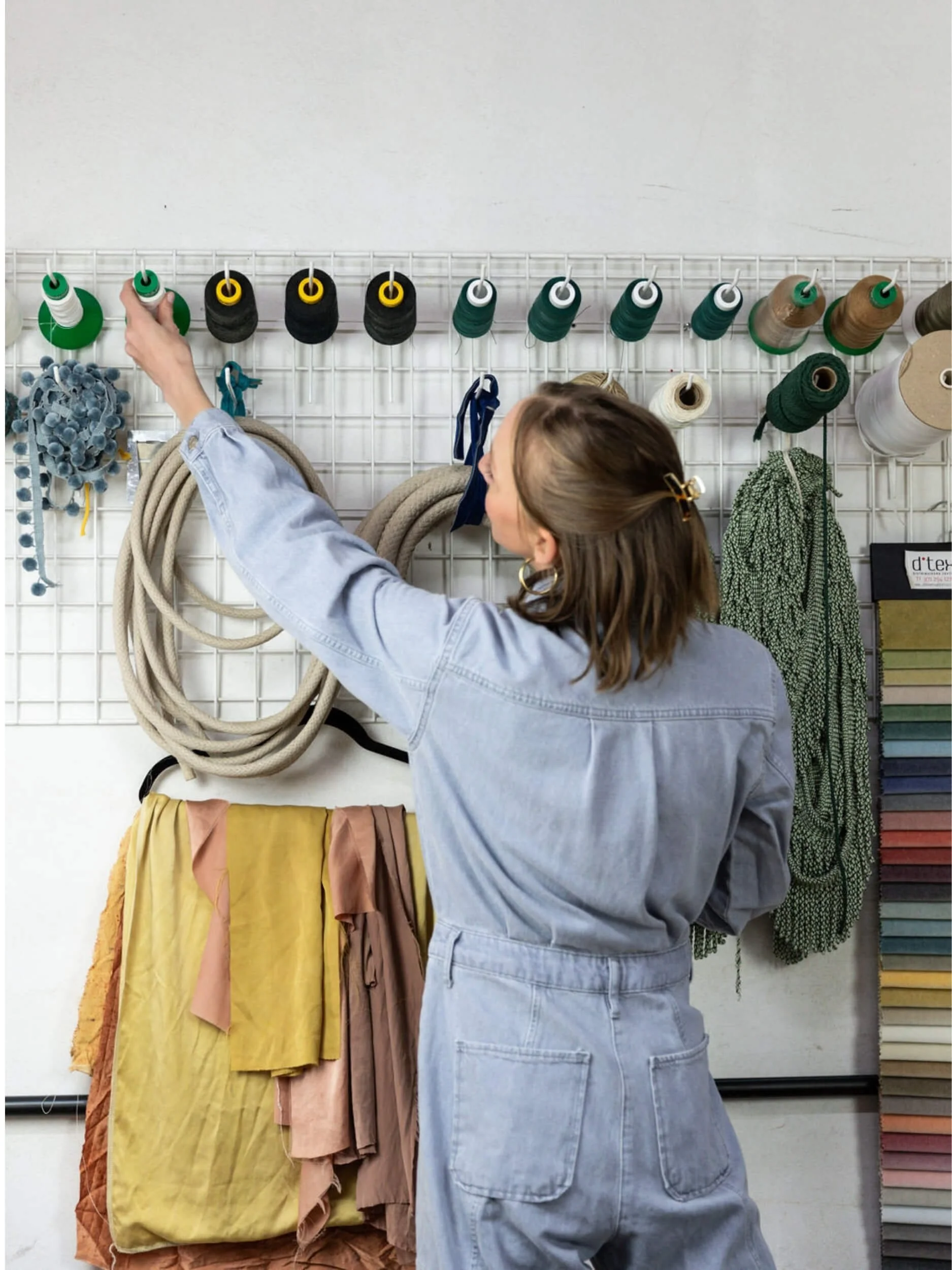 A person with brown hair and gold hoop earrings wearing a light blue denim jacket is selecting thread spools from a display on a white grid wall, with various colored fabrics hanging below.