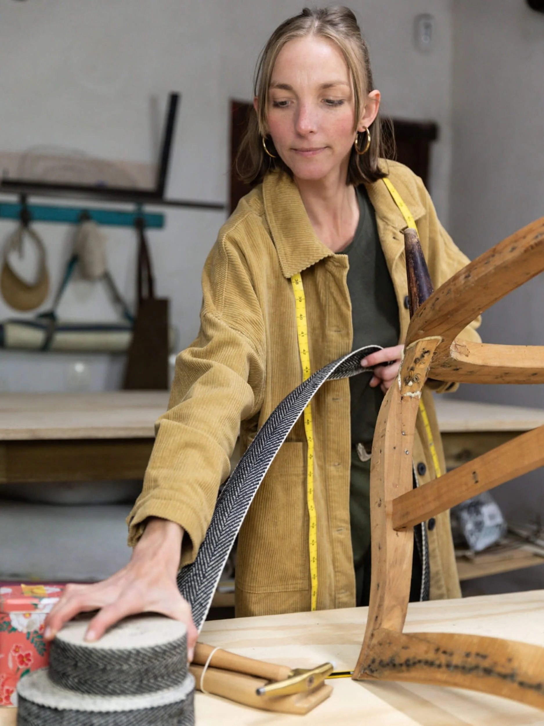 A woman with earrings and a yellow jacket is working on a woodworking project, measuring a wooden chair with a measuring tape.