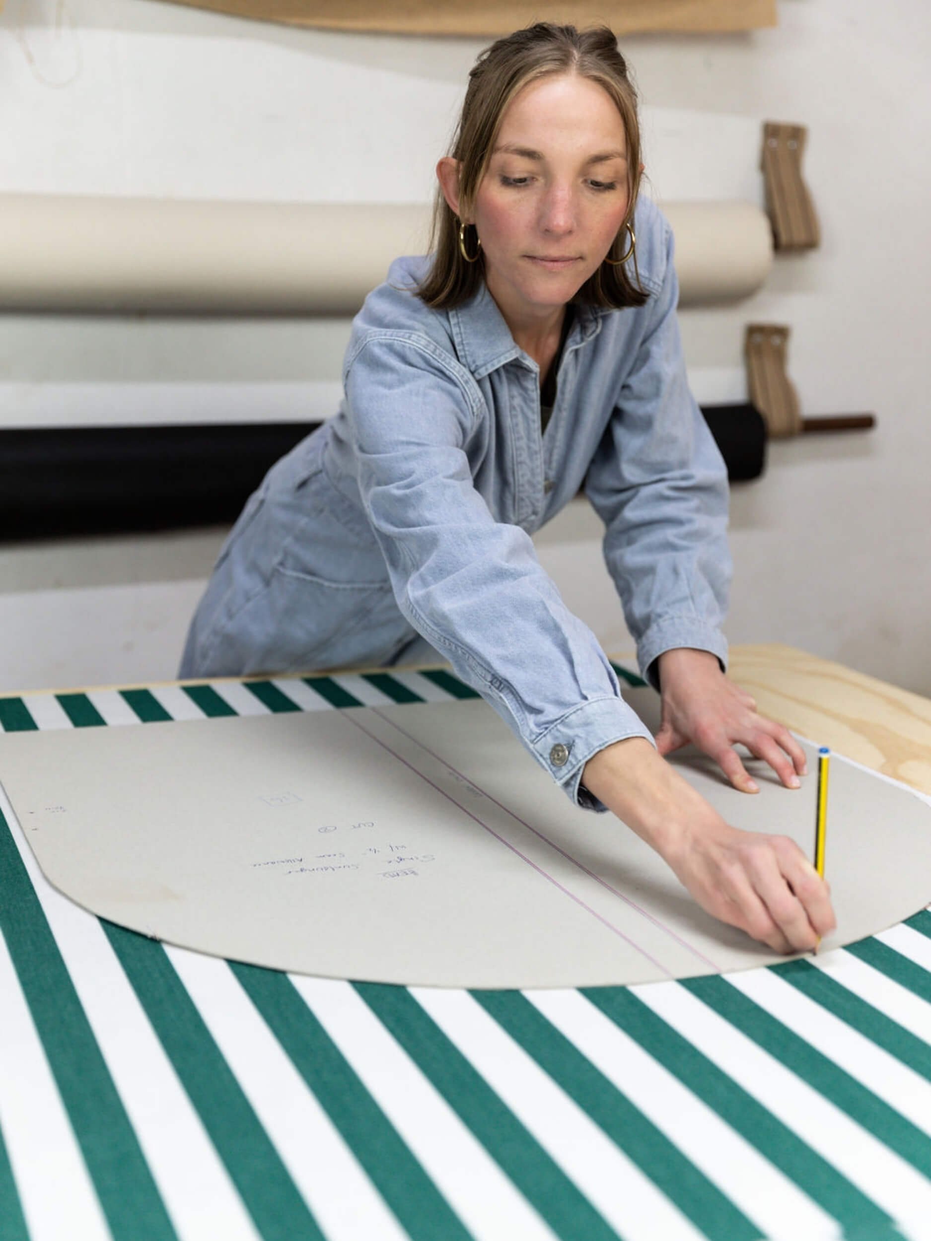 A woman is working with a large sheet of paper on a table with a green and white striped border. She is using a yellow and black tool, possibly a measuring or cutting device, and appears to be marking or measuring the paper.