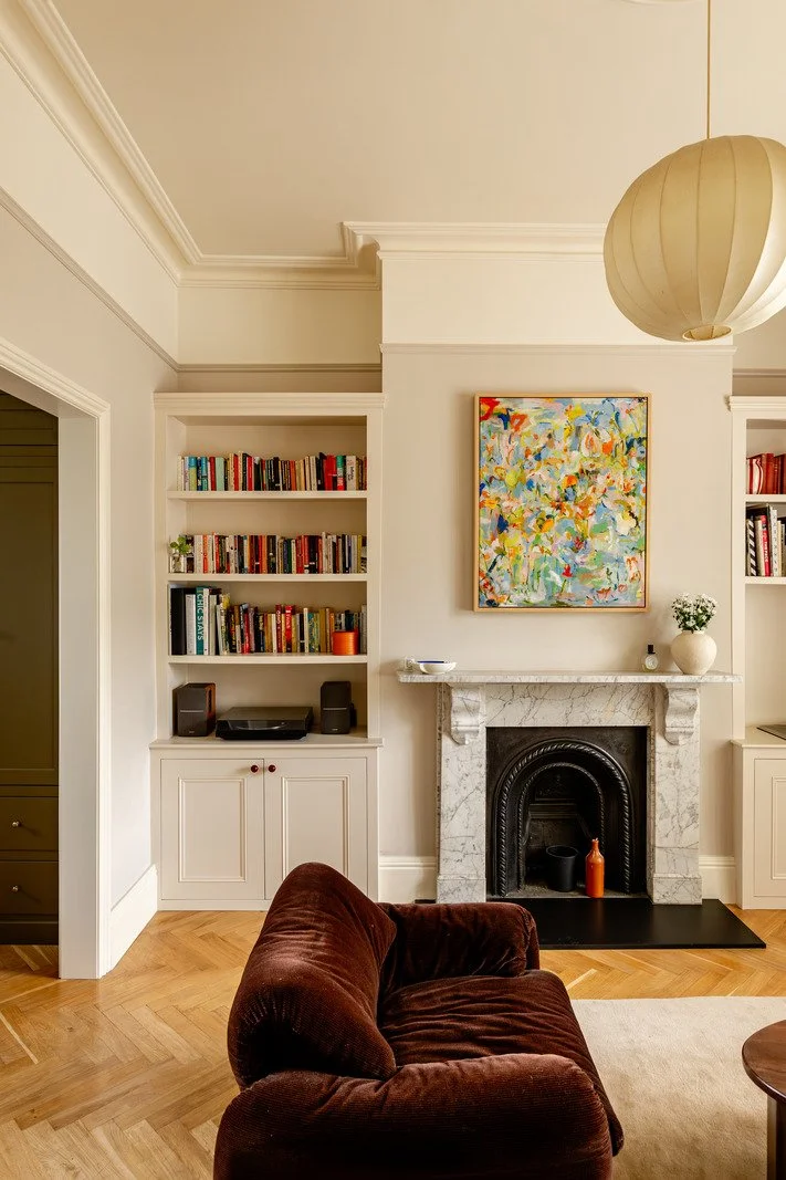 Living room with built-in white bookshelves on either side of a marble fireplace, a colorful abstract painting above the fireplace, a round brown velvet armchair in the foreground, and a large paper lantern ceiling light.