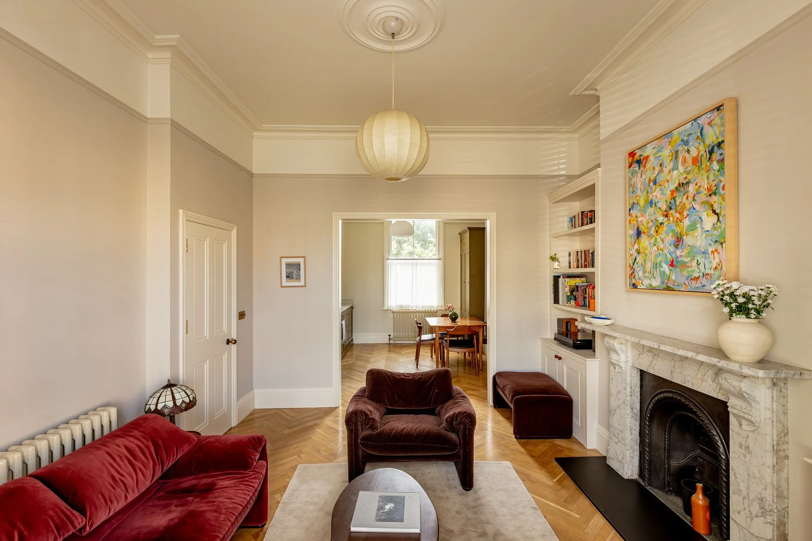Living room with fireplace, colorful abstract painting, brown and red velvet chairs, white built-in bookshelves, and a doorway leading to a dining area with a window, wooden dining table, and chairs.