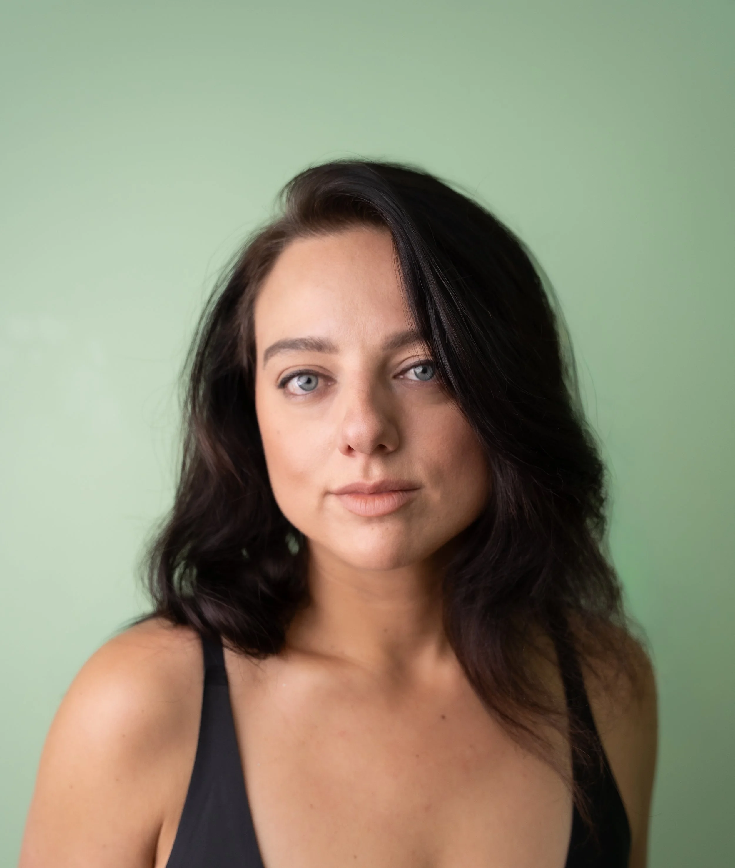 Close-up portrait of a woman with black hair, blue eyes, and fair skin wearing a black top against a light green background.